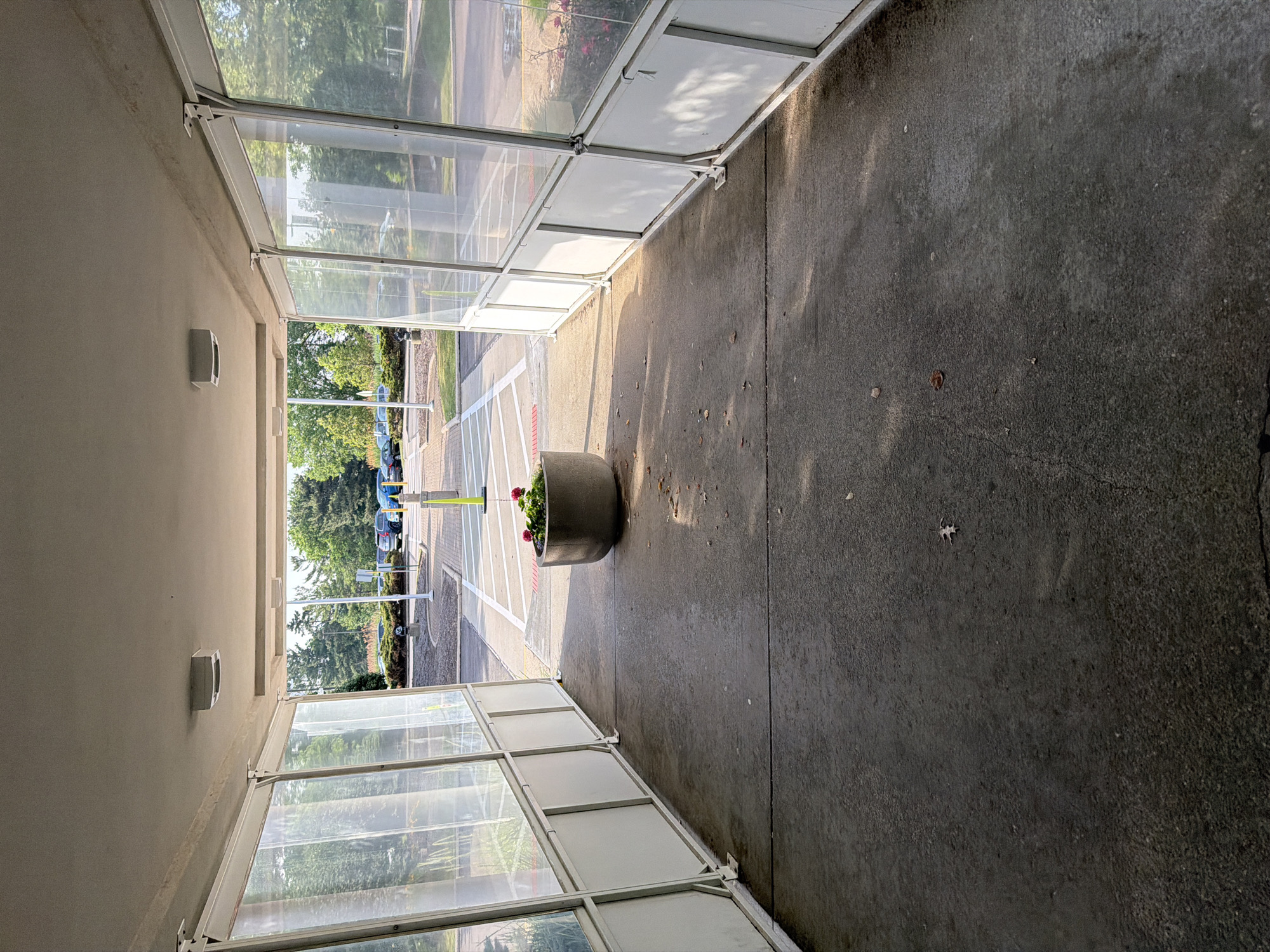 The image shows a covered walkway or tunnel with clear plastic siding on both sides, leading to an outdoor area with a parking lot. The walkway has a concrete floor and a white metal frame. Two fluorescent light fixtures are mounted on the ceiling of the walkway. In the center of the walkway, a round concrete planter with red flowers is visible. Beyond the walkway, a parking lot with several cars and trees can be seen. The scene appears to be in Beachwood, United States of America. The lighting suggests it is daytime, with sunlight casting shadows on the walkway floor.