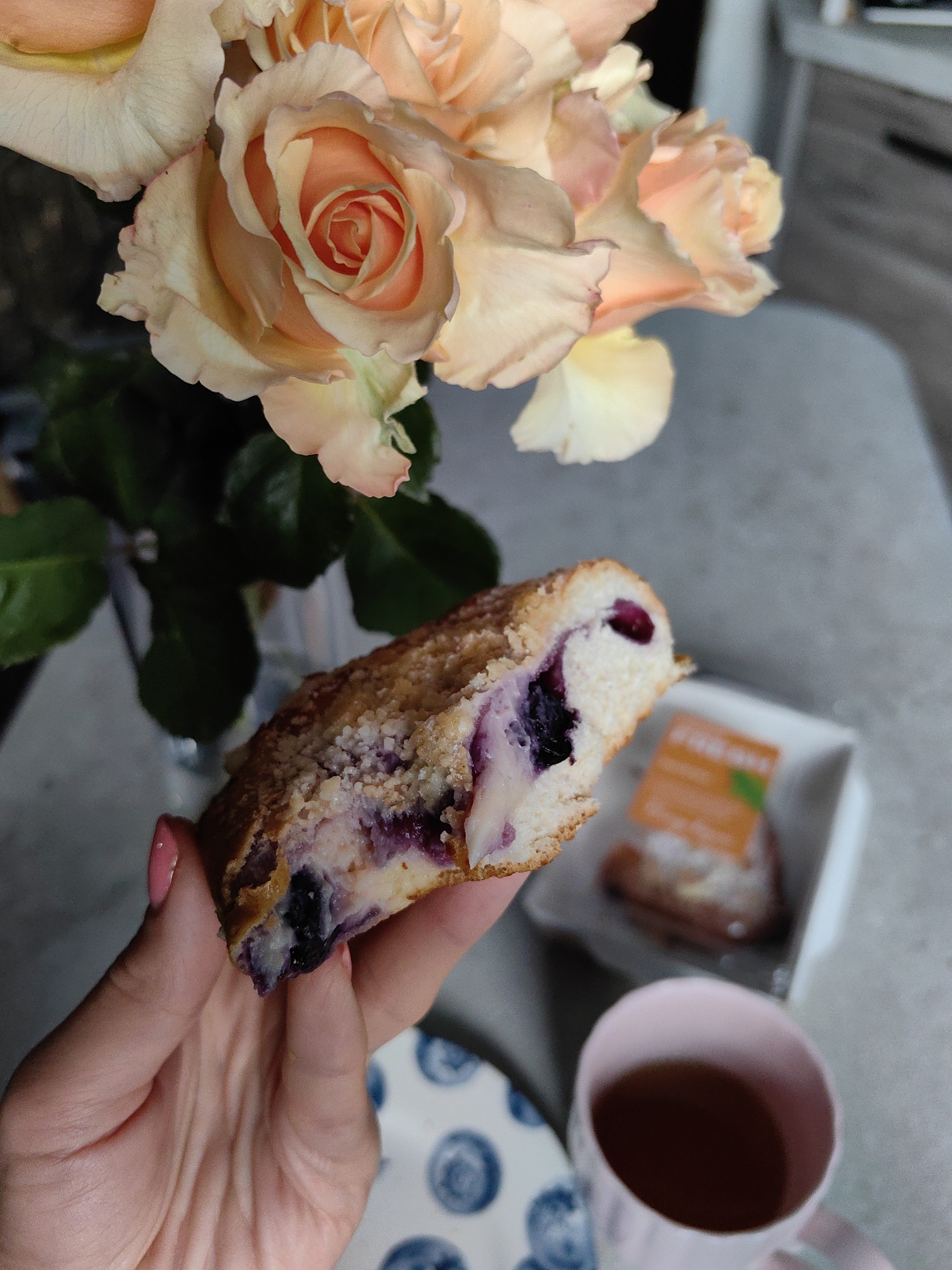 A close-up shot shows a hand holding a slice of blueberry coffee cake. The cake has a crumb topping and visible blueberries within the batter. In the background, a bouquet of peach-colored roses is partially visible. To the right, a small white dish containing another piece of cake or pastry sits on a grey countertop. A pink mug filled with a dark liquid, likely tea or coffee, is also present. A patterned blue and white plate is seen at the bottom left. The setting appears to be indoors, possibly a kitchen or dining area. There is no indication of the location being in the city of Fanipal, Belarus.