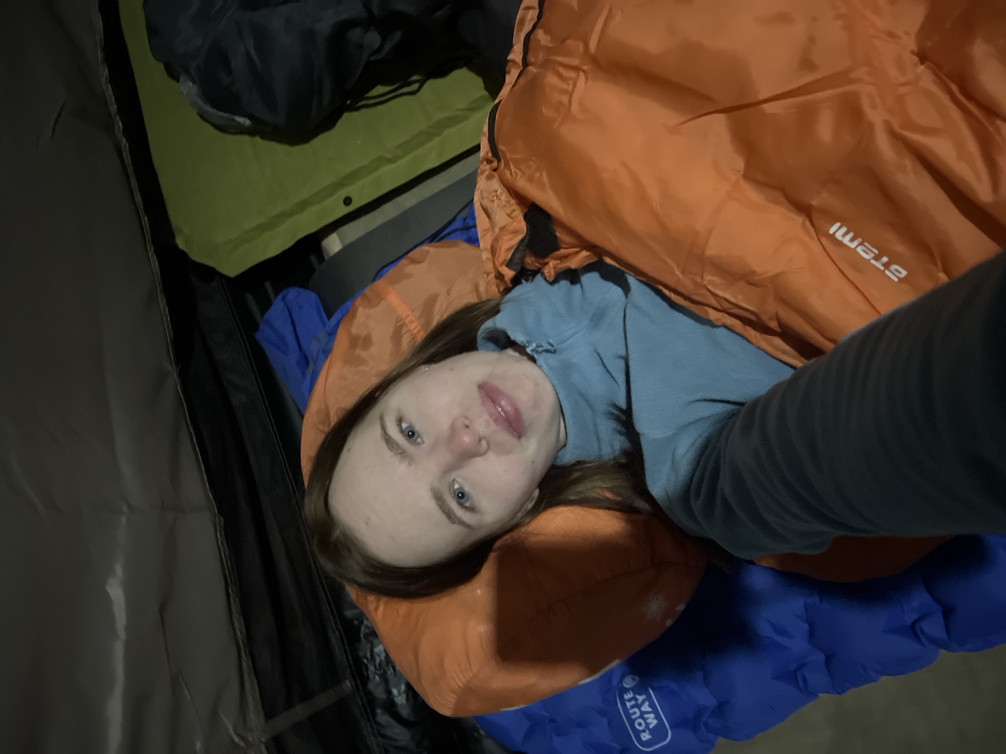 This media file captures a close-up, upward-angled selfie of a young woman resting inside a tent or similar outdoor shelter, likely during a camping trip in Lesnoye, Russia. She has long brown hair, blue eyes, and fair skin, looking directly at the camera with a calm, pleasant expression and a slight smile.
She is wearing a light blue long-sleeved top and appears to be comfortably situated for sleep or rest. Her head rests on an orange pillow, and she is lying on a blue inflatable sleeping pad, which has the mirrored text "ROUTE WAY" visible. An orange sleeping bag or top quilt covers her, with the mirrored brand name "STEMI" printed on it. The selfie perspective accounts for the reversed text on the items.
The dim, enclosed lighting suggests it is either night or early morning within the tent. Various pieces of dark camping equipment are visible in the background, including dark green fabric, possibly a tent floor or ground tarp, and dark grey/black fabric from the tent structure or other gear. A dark jacket or bag is also partially seen in the upper left corner. The scene conveys a sense of comfort and relaxation within a rugged outdoor setting.