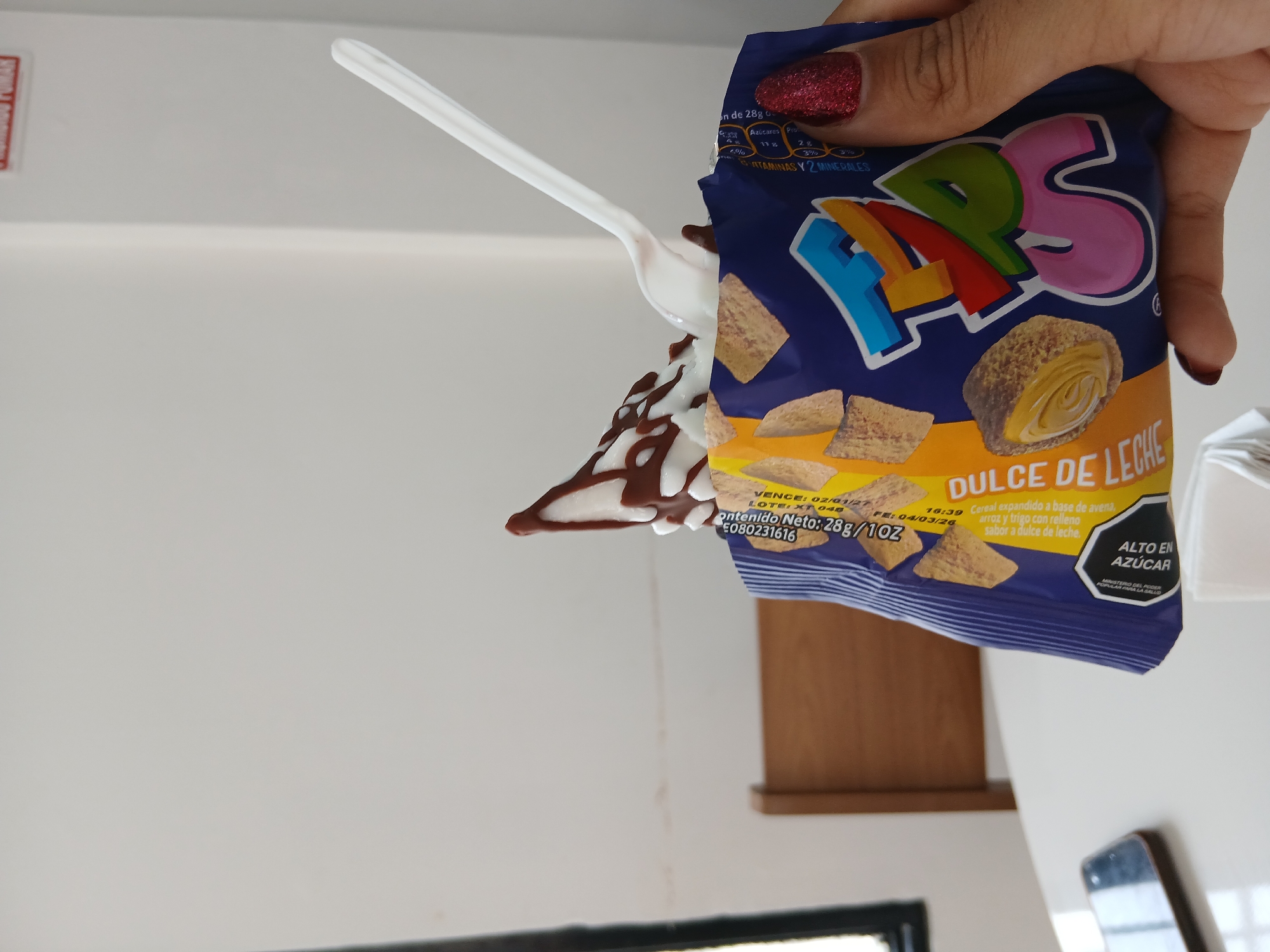 The image shows a hand holding an open bag of "Flaps" Dulce de Leche cereal. A white plastic spoon with a dollop of ice cream covered in chocolate syrup is emerging from the bag. The bag is predominantly blue, with the "Flaps" logo in colorful bubble letters at the top. The packaging indicates "Dulce de Leche" flavor, and includes a warning about high sugar content. The hand holding the bag has red glitter nail polish. The background appears to be a wall and a counter. There is no explicit indication of Juan Griego, Venezuela in the image.