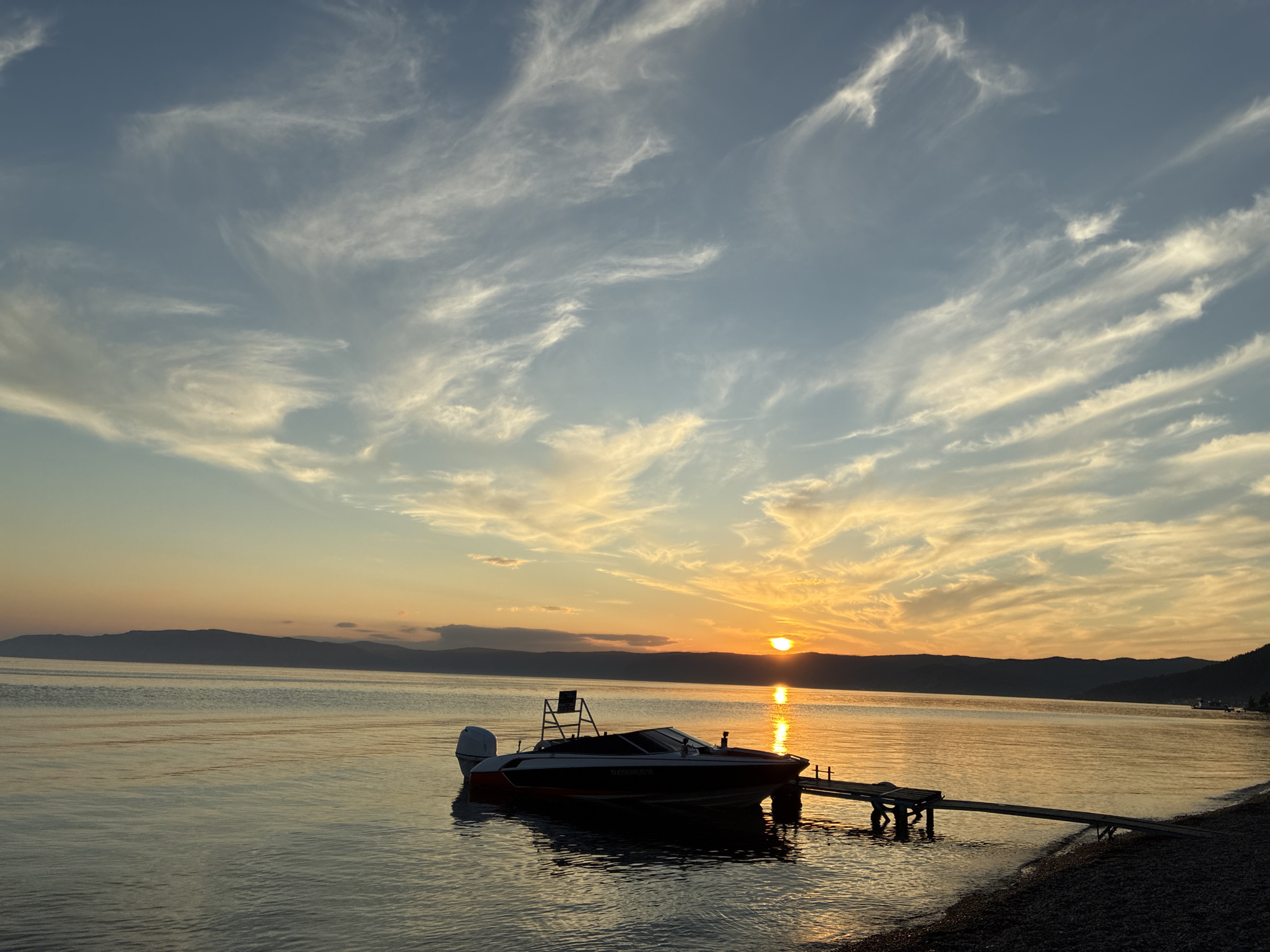 A scenic sunset over Lake Baikal in Listvyanka, Russia, is captured in this wide-angle shot. The sun, a brilliant orange orb, is setting behind a silhouetted mountain range in the distance, casting a warm glow across the water and sky. Wispy, white clouds are scattered across the blue sky, reflecting the golden hues of the sunset.
In the foreground, a small motorboat, red and white with a black hull, is moored to a wooden pier extending from the pebbled shoreline. The boat is dark and appears to be unoccupied. The water is calm, with gentle ripples reflecting the colors of the sky. The shoreline is composed of small, dark pebbles. The overall mood of the image is serene and peaceful, conveying the beauty of a natural landscape at dusk.