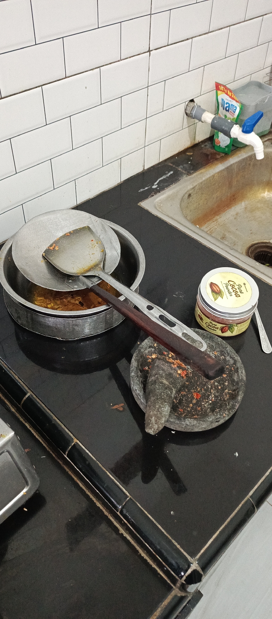 The image depicts a kitchen counter scene in what appears to be a residential kitchen in Arjasa, Indonesia. The counter is black and glossy, with a tiled backsplash composed of white subway tiles. To the right, a stainless steel sink is visible, showing signs of rust and use. A faucet with a blue handle is attached to the wall above the sink, and a package of "Mama" brand dish soap is placed nearby.
In the foreground and mid-ground of the image, several kitchen implements are arranged on the counter. A stainless steel pot with a mesh sieve placed on top contains some yellow, possibly food-related, material. A metal spatula with a wooden handle rests on the sieve and the pot's rim. To the right of the pot, a granite mortar and pestle are present. The mortar contains red chili fragments, indicating recent use for grinding spices. A small jar labeled "Pure Cocoa Powder" is also visible near the mortar and pestle. The overall impression is of a working kitchen, possibly mid-meal preparation or cleanup.