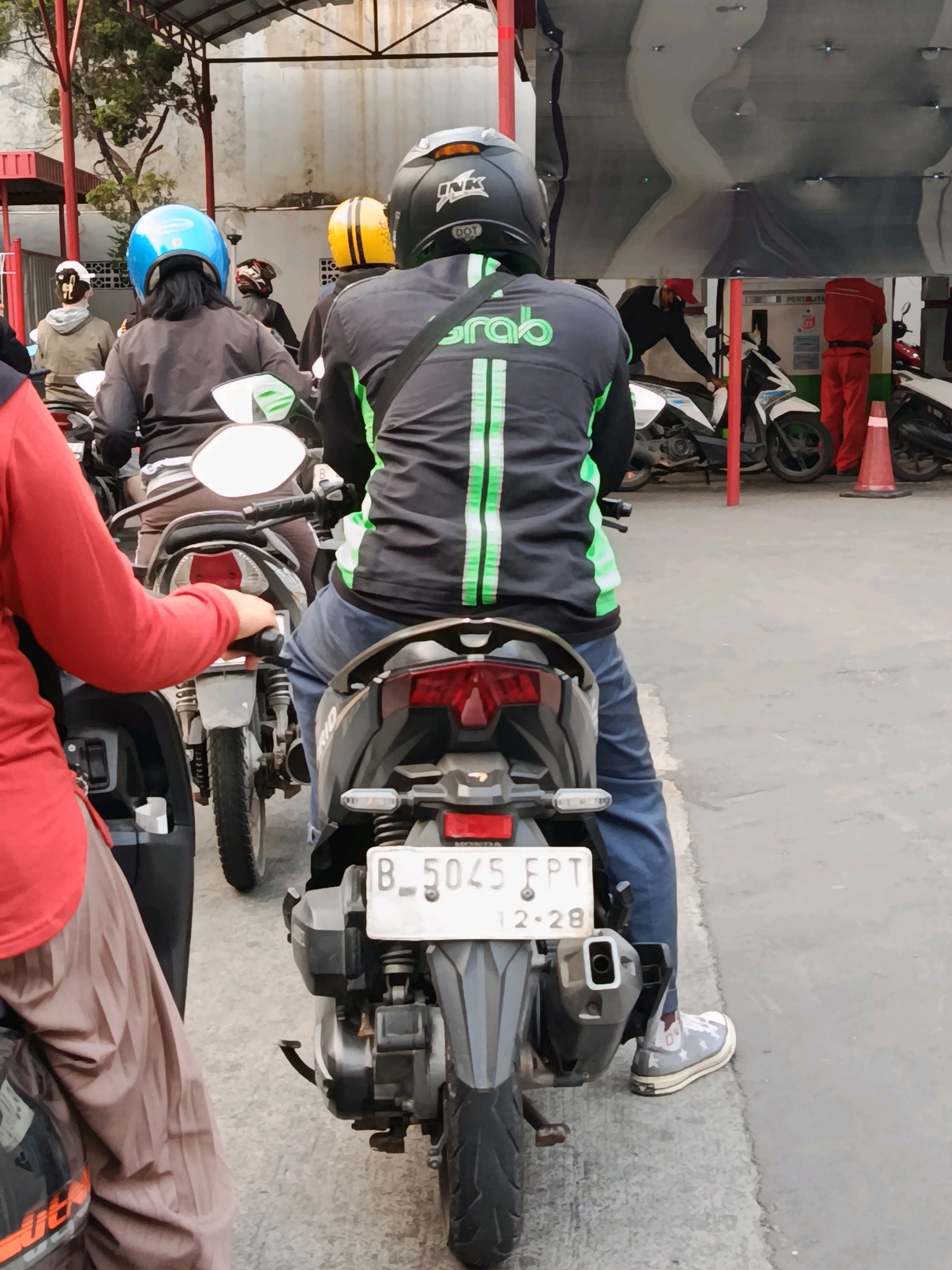 The image depicts a street scene in Bekasi, Indonesia, likely at a gas station given the presence of pumps in the background. The foreground is dominated by several individuals on motorcycles. The primary subject is a person wearing a dark jacket with green vertical stripes and a black helmet, sitting on a motorcycle and facing away from the camera. This individual is wearing blue jeans and sneakers with a star pattern. To the left, another person in a red shirt and light brown pants is partially visible, also on a motorcycle. Further back, other motorcyclists are seen, including one wearing a blue helmet and another with a yellow and black helmet. The environment appears to be outdoors with a paved ground surface and a covered area overhead, supported by poles. The overall atmosphere suggests a queue or waiting area, possibly for fuel.