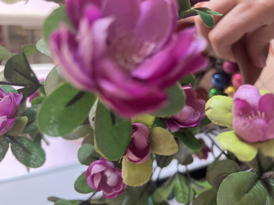 The scene depicts a close-up, shallow-depth-of-field view of artificial floral arrangements within an indoor setting in Кез, Russia. The foreground is dominated by a large, heavily blurred magenta-purple artificial flower, with its petals and a lighter center vaguely visible. Throughout the mid-ground, numerous smaller artificial flowers with magenta-purple petals and green fabric leaves are arranged on stems. Some leaves are a lighter, yellowish-green hue. Several of these smaller flowers appear partially open or in bud form. In the lower right background, a light-skinned human hand, possibly belonging to an adult, is visible, with fingers slightly curled, suggesting an interaction with objects beyond the immediate floral arrangement. Below the flowers and near the hand, a collection of small, reflective, spherical objects in various colors including blue, green, pink, and gold are observed, likely contained within a receptacle on a surface. The background is brightly lit and heavily blurred, consistent with an interior space.
