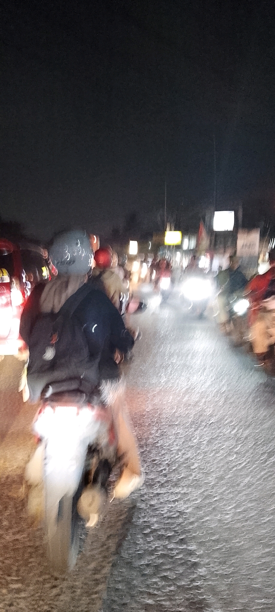The image depicts a nighttime scene on a street in Depok, Indonesia, characterized by motion blur indicative of a moving vehicle. In the foreground, a person wearing a helmet and a dark jacket is riding a motorcycle, illuminated by the headlights of other vehicles. The road surface appears wet or reflective, with numerous streaks of light suggesting movement. The background is indistinct due to the blur but shows the lights of other motorcycles and buildings, with some signs and a flag visible. The overall impression is of a busy street at night.