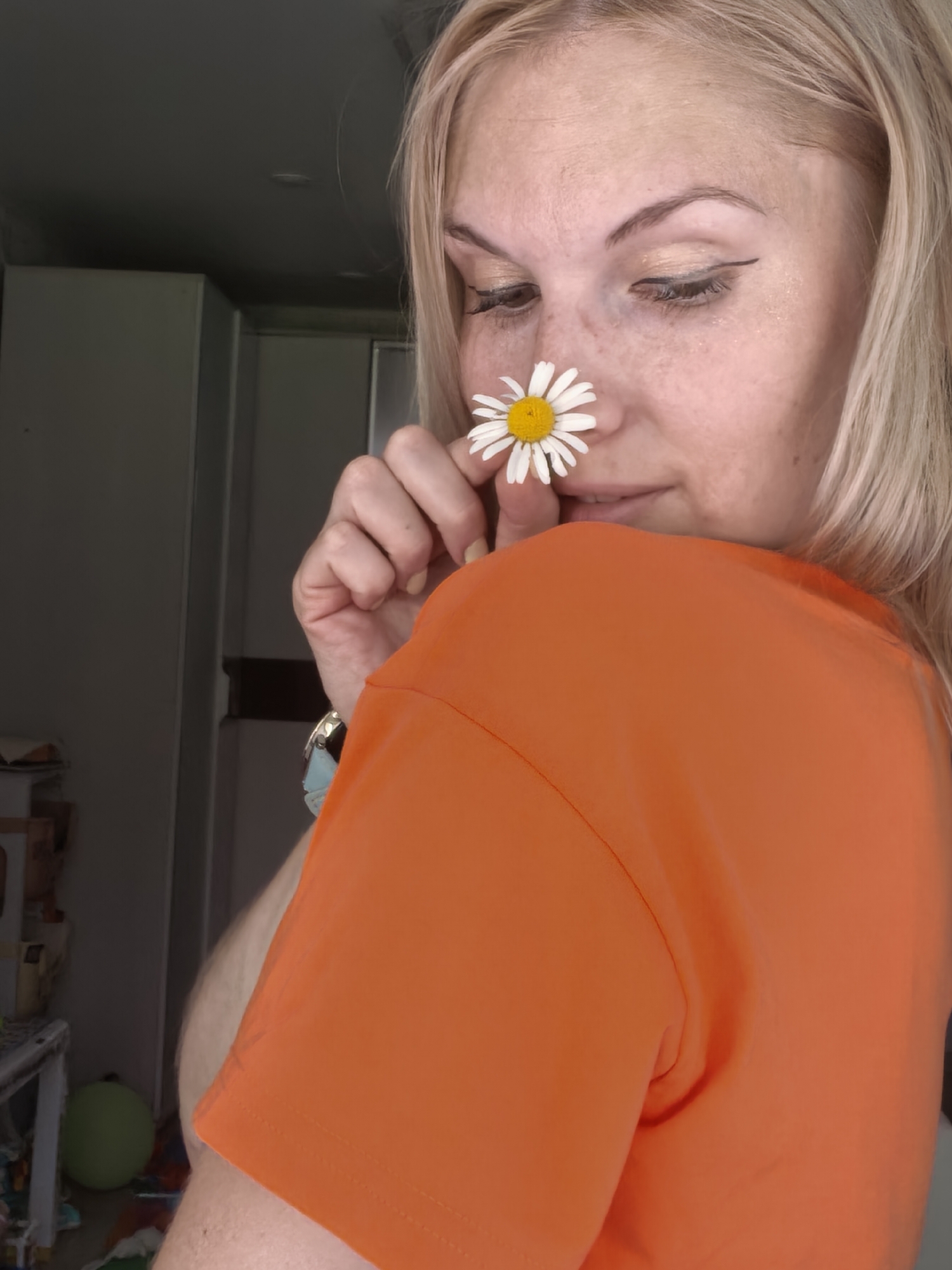 A blonde woman in an orange t-shirt is holding a white daisy up to her nose, as if to smell it. She is looking down and to her left. The background is a blurry interior with gray cabinets and a dark ceiling. There is a hint of what appears to be a kitchen or laundry room. The image does not provide any specific information about the city of Sterlitamak, Russia.