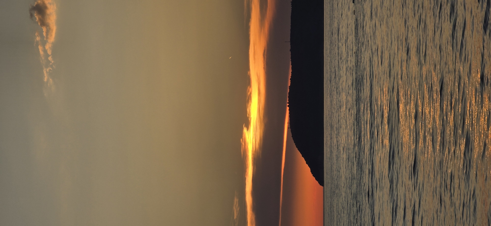 The image depicts a sunset over the Adriatic Sea, possibly near Bar, Montenegro. A dark silhouette of a landmass, likely a mountain or coastline, divides the scene. To the left, the sky is painted with hues of orange and yellow, indicating the setting sun. The sun's rays reflect off the water's surface on the right, creating shimmering patterns. The water is relatively calm with gentle ripples. There are no visible people or other specific objects like boats.