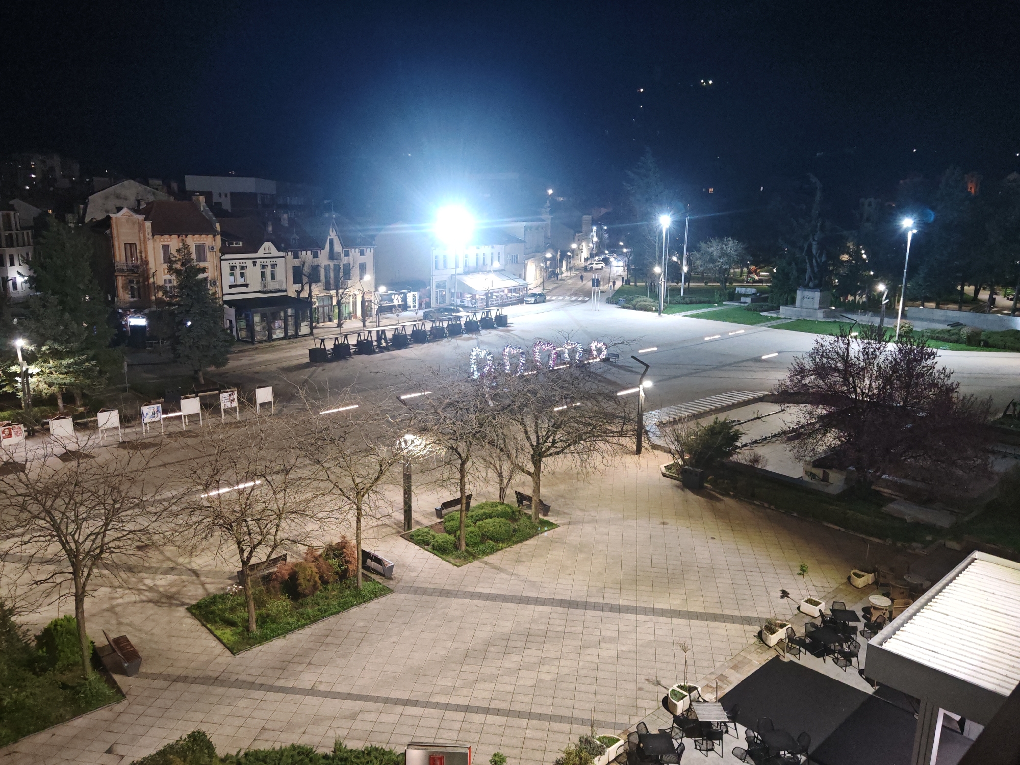 The image captures a nighttime scene of a public square in Vratsa, Bulgaria. Dominating the view are expanses of light gray paving stones, patterned with dark gray lines. The square is illuminated by numerous streetlights, which cast a bright glow and long shadows.
Several deciduous trees with bare branches are scattered throughout the square, some enclosed in rectangular planters with green bushes. A set of benches are placed around the trees.
In the background, a row of buildings lines the square. Their architecture is a mix of styles, with some featuring gabled roofs and others flat facades. The buildings are illuminated by the streetlights and lights from within.
In the distance, there are streetlights and cars parked along the street. The night sky is dark.
There are several sets of tables and chairs, that are likely part of an outdoor cafe or restaurant setting. The tables are empty. A partially visible roof of a building is at the bottom right corner of the image.