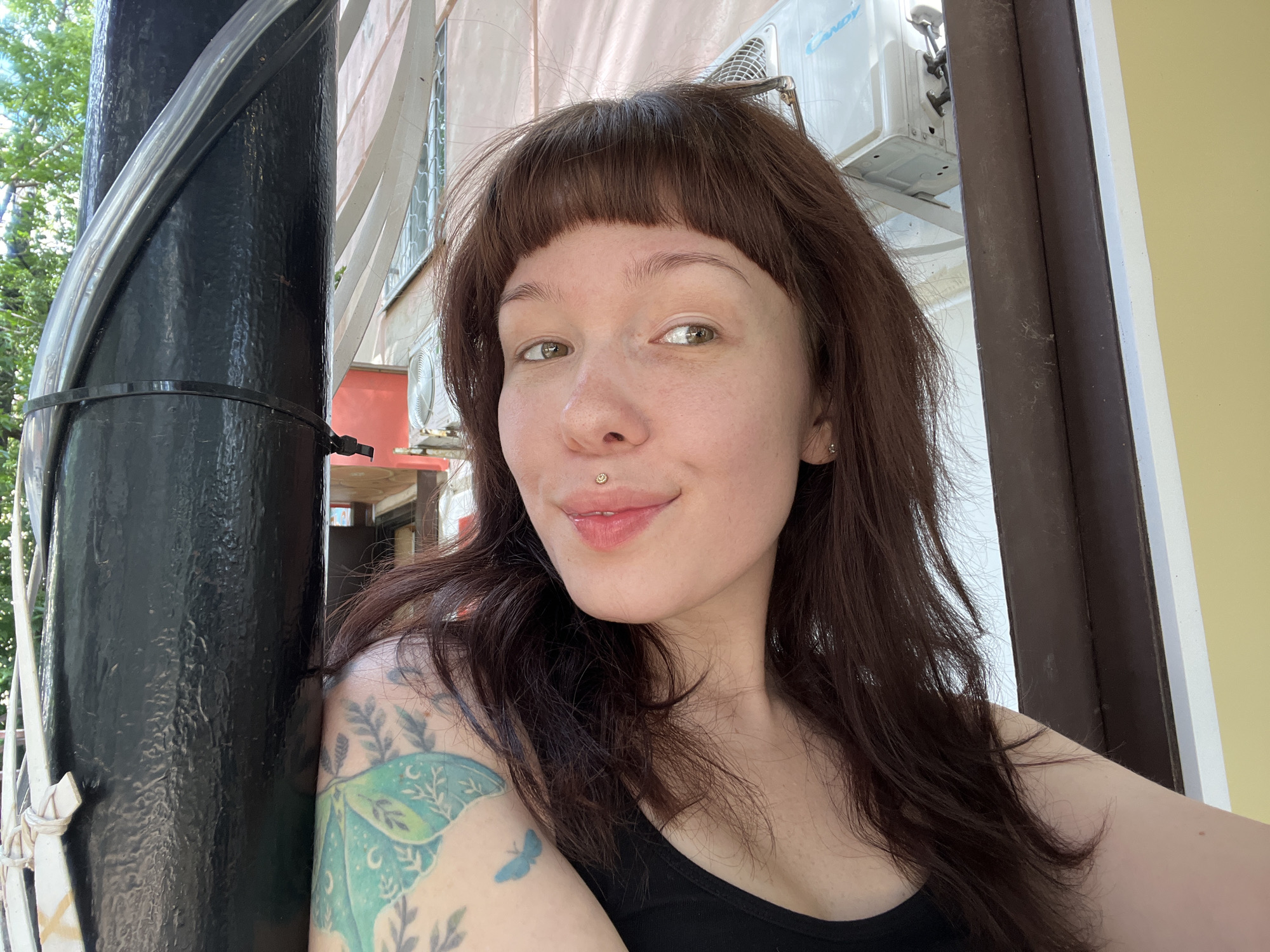 A young woman with brown hair and bangs, wearing a black tank top, takes a selfie outdoors. She has a lip piercing and a tattoo on her left arm. She is leaning against a black pole that has a cable wrapped around it. In the background, there is a building with an air conditioning unit visible. The scene appears to be in a city setting.