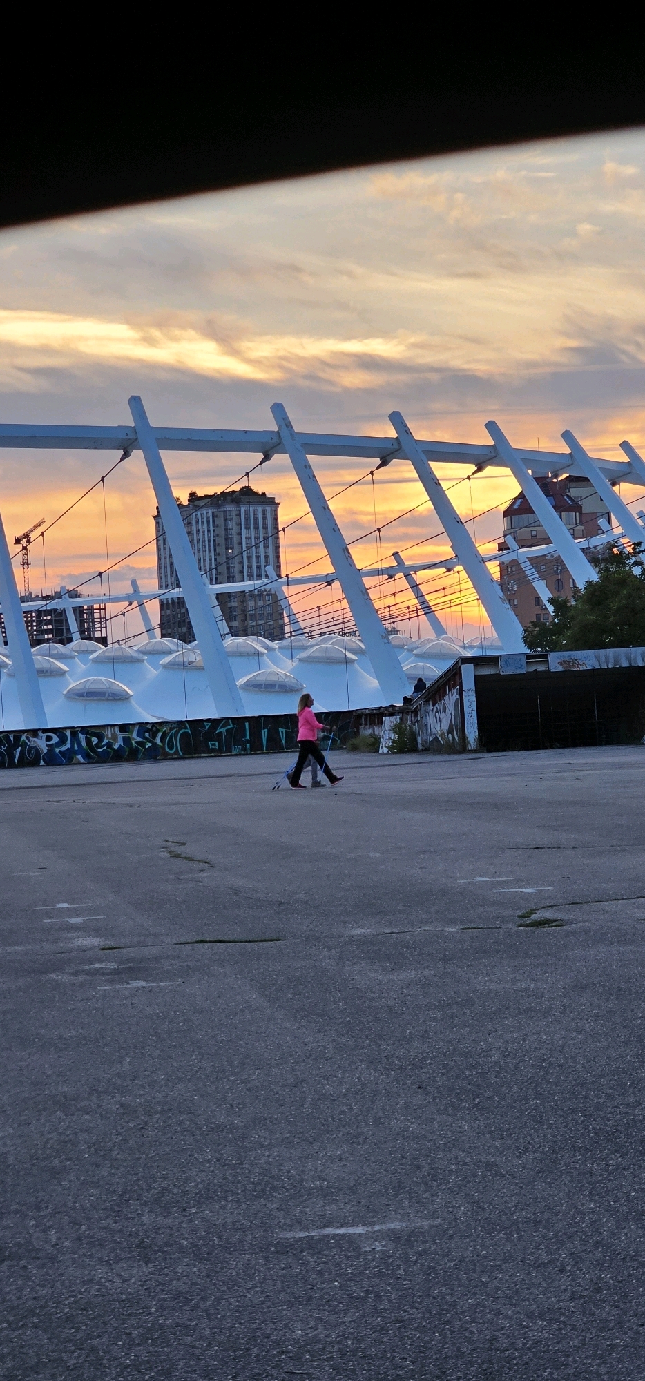 A medium shot shows a woman with blonde hair, wearing a bright pink long-sleeved shirt and black pants, engaged in Nordic walking across a paved area. She holds two Nordic walking poles, one in each hand, and moves from left to right across the frame. The background features the distinctive white, angled supports of a stadium structure, with a cloudy sky painted in hues of orange, yellow, and pink, indicative of sunset. Underneath the stadium structure, the white, domed roofs of what appear to be stadium seating are visible, adorned with graffiti. Beyond the stadium, a tall, light-colored building stands out, and to its left, a construction crane is visible. The ground is a weathered, gray asphalt surface with some cracks and minor debris.