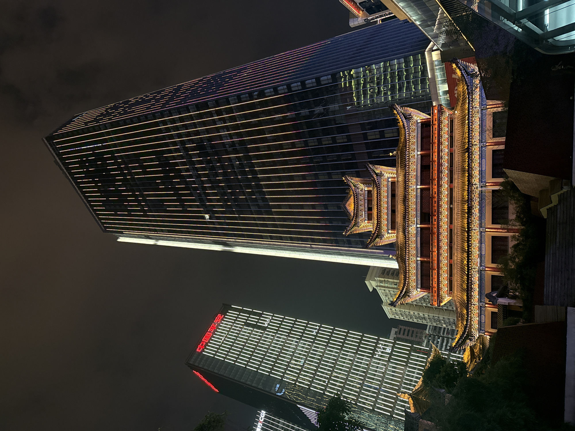 At night in Yuzhong District, China, the image features a juxtaposition of traditional and modern architecture. Two modern skyscrapers are visible, illuminated with horizontal lines of white and amber light. One building has red characters on its side. Adjacent to these, a traditional Chinese building with multiple tiers and ornate, brightly lit features stands out. The sky is dark and overcast, suggesting it is nighttime. The composition is vertical.