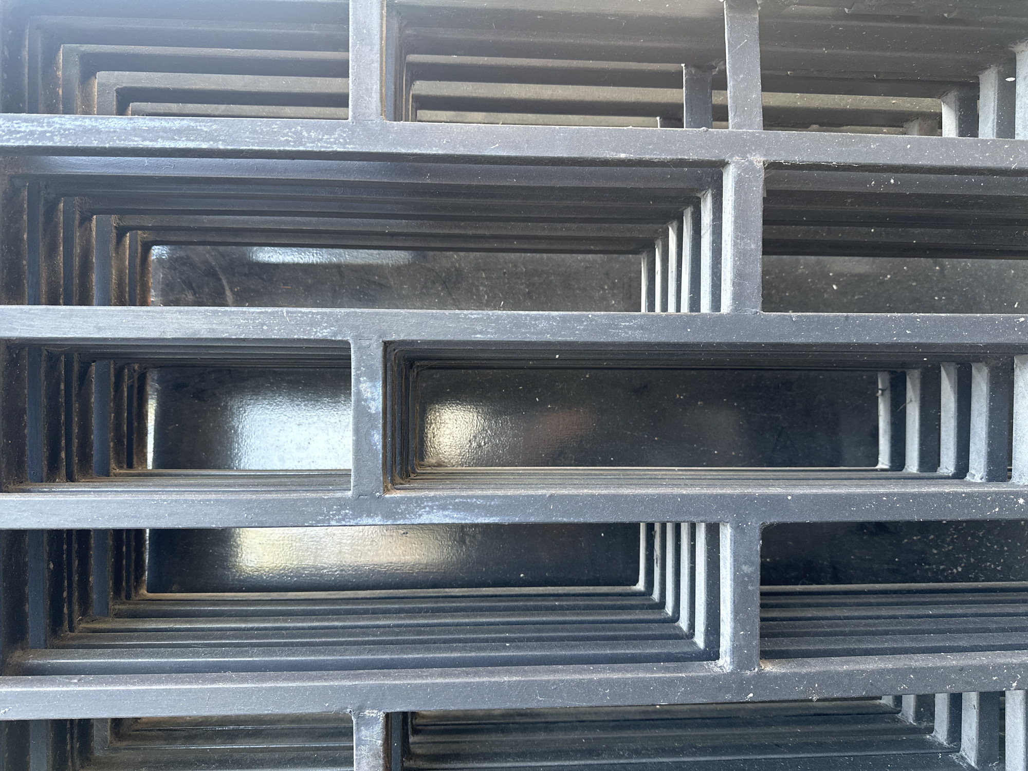 This is a close-up, eye-level photograph of a stack of black metal grates. The grates are rectangular and nested inside each other, creating a sense of depth and repetition. The metal appears to be painted and shows some signs of wear, with small white specks scattered across its surface. The background behind the grates is dark and reflective, suggesting a wall or another dark surface. The lighting appears to be natural, casting subtle shadows and highlights on the metal. There are no people or animals visible in the image. The scene is forensic and matter-of-fact, documenting the object without any implied narrative. The location in Parangjoro, Indonesia is noted, but the image itself does not provide visual cues to confirm this specific location; it depicts a generic industrial or storage setting.