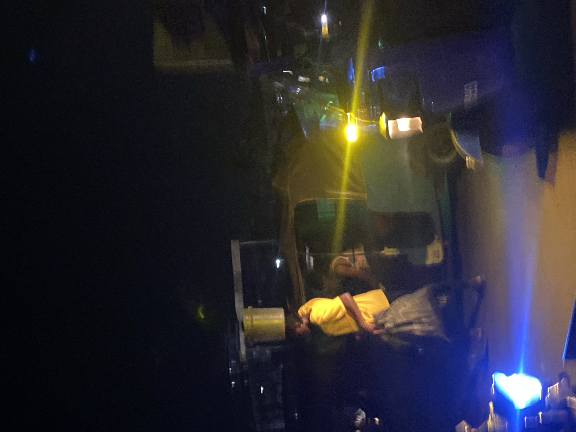 At night in Abakaliki, Nigeria, a person wearing a bright yellow shirt is walking with a bucket balanced on their head and carrying a large bag in their arms. They are on a street with vehicles, including an auto-rickshaw. The scene is illuminated by streetlights and vehicle headlights, creating strong yellow and blue light flares. The background is dark, suggesting an urban environment at night.