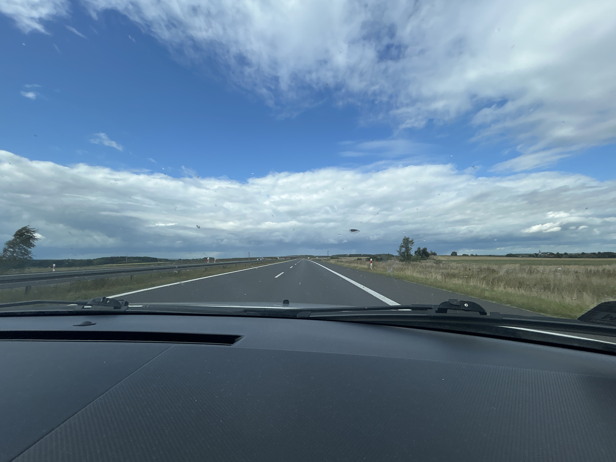 The image is taken from the interior of a car, looking out through the windshield. It shows a highway with two lanes in each direction, separated by a median. The road is asphalt and has white lane markings. On either side of the highway, there are fields of dry grass and scattered trees. In the distance, there are hills and a cloudy sky. The windshield has several water droplets and smudges on it. The dashboard of the car is visible in the foreground. The location is identified as Witosław, Poland.