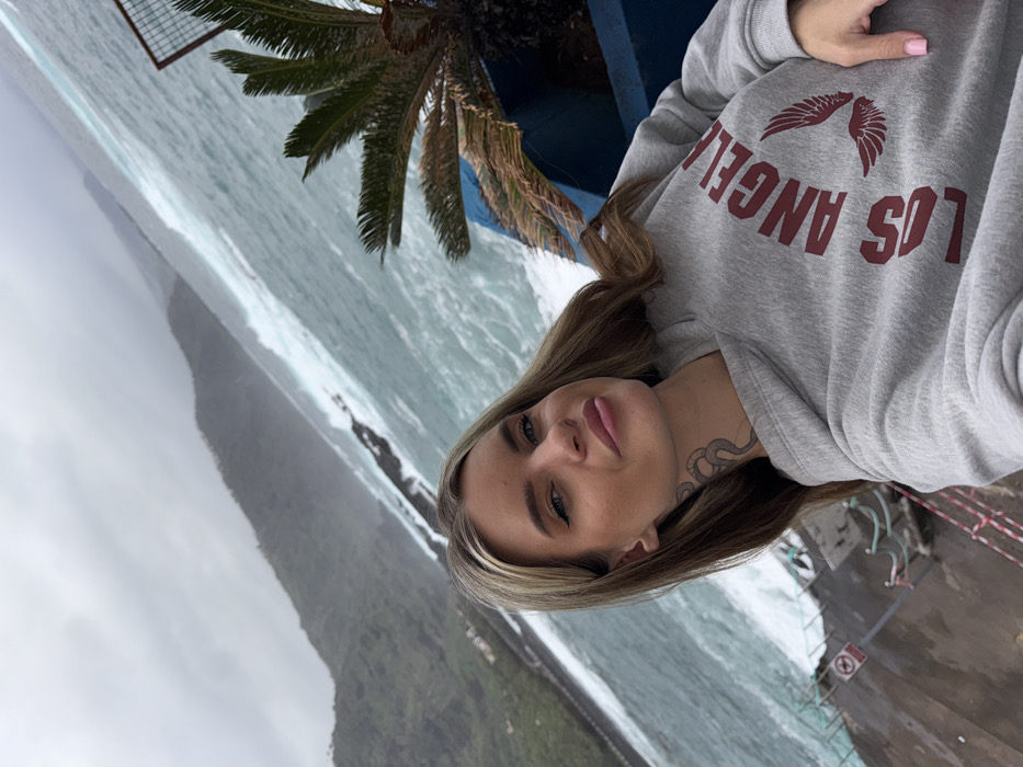 A woman poses in a grey "Los Angeles" sweatshirt. She has blonde hair and a snake tattoo on her neck. The background features a choppy ocean, a green, mountainous landscape obscured by clouds, and a palm tree. A metal barrier is visible in the upper left corner. The photo appears to be taken from a high vantage point.