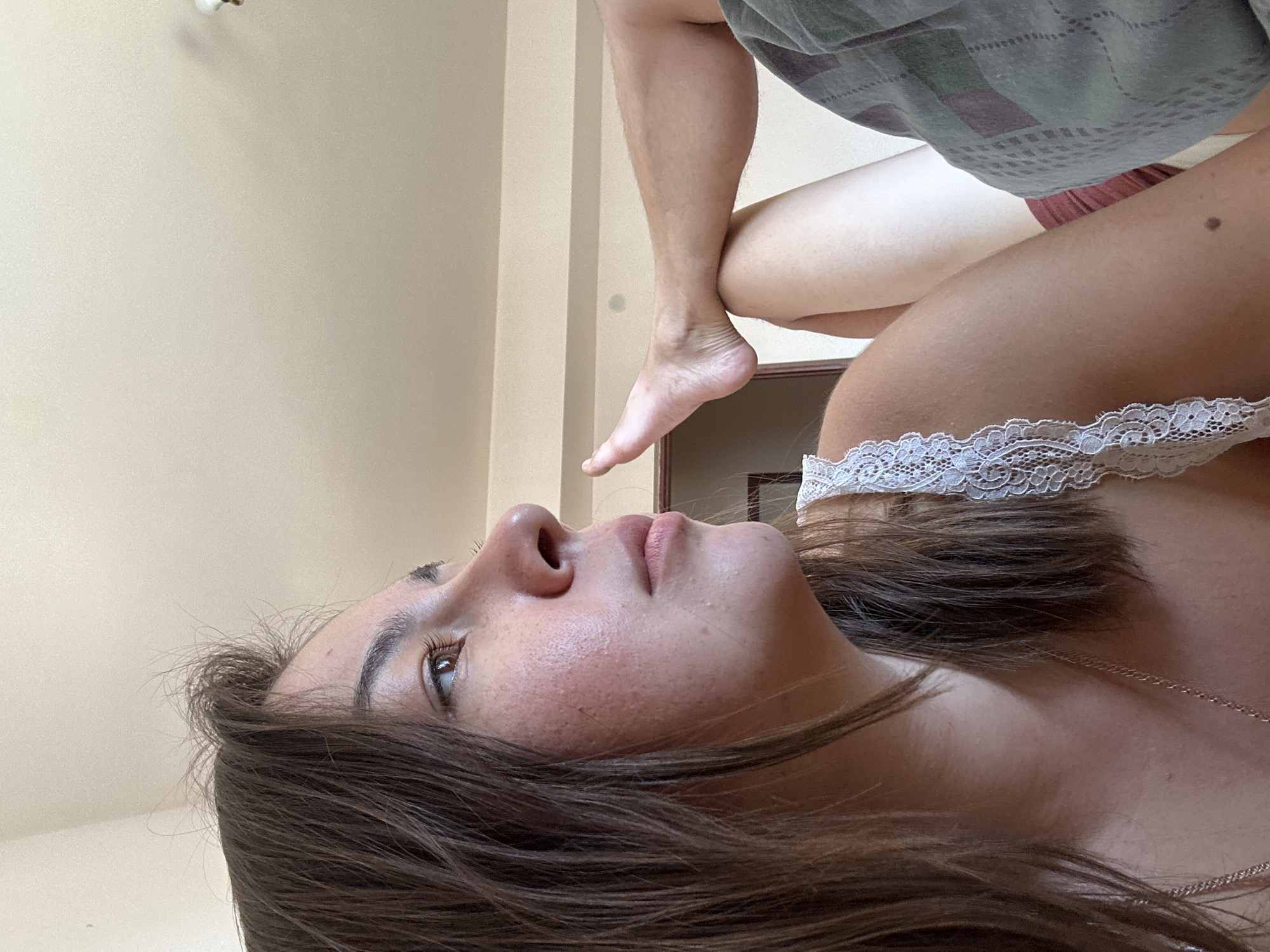 The image shows a close-up, side profile of a young woman's face. She has dark brown hair, which is slightly messy, and her eyes are looking to her left. Her skin appears fair with some visible pores and blemishes. She is wearing a white lace-trimmed top. Her left arm is bent at the elbow, and her hand is resting on her leg, which is visible in the lower right portion of the image. The leg is clad in a patterned garment. The background is a neutral-colored wall and ceiling. There is no indication of the location being the city of Gyumri, Armenia.