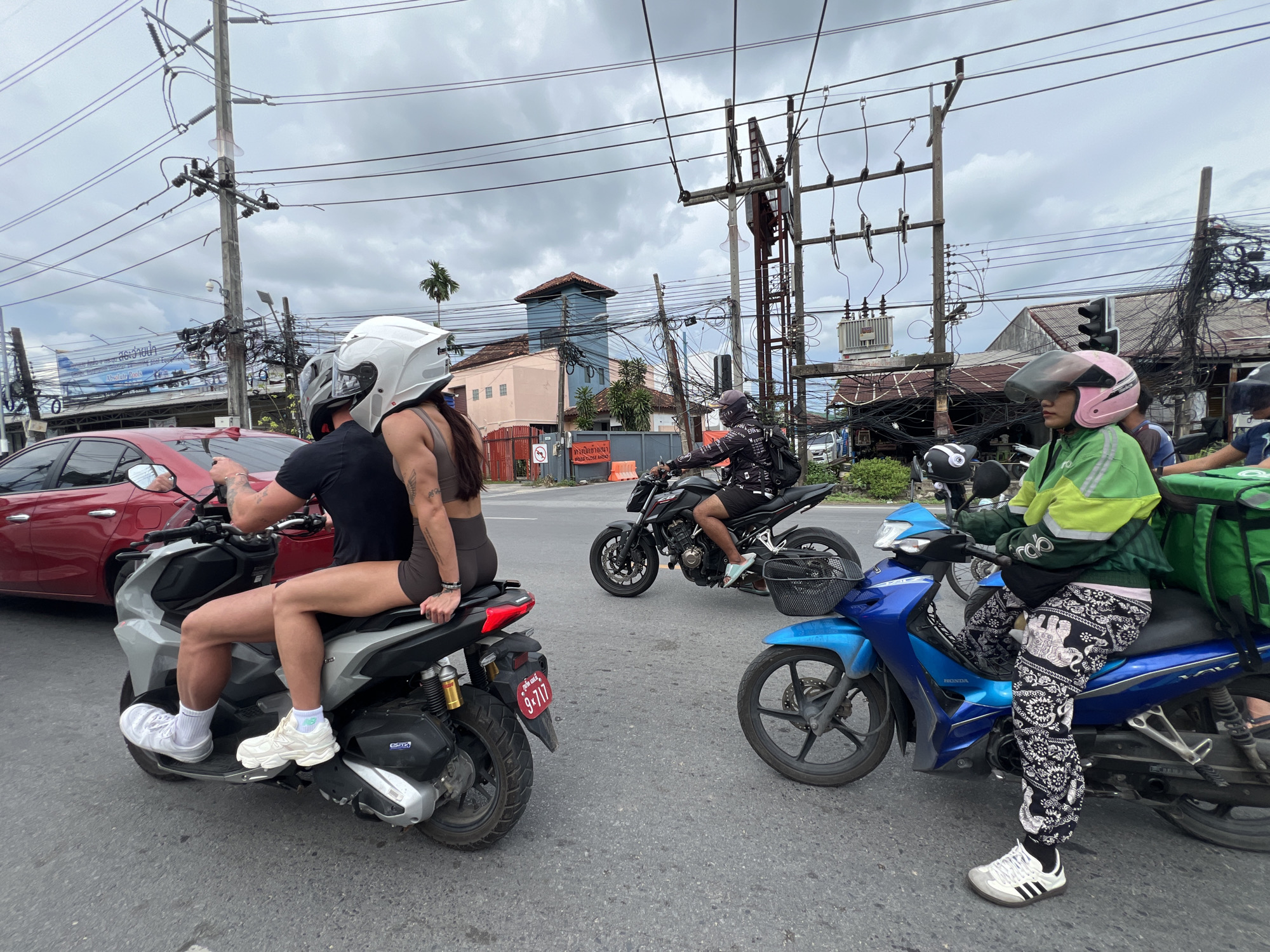 In Chalong, Thailand, on a cloudy day, three motorcycles are stopped at an intersection. A red car is parked on the left side of the frame, behind a silver scooter occupied by a man and a woman. The woman, wearing a white helmet and a dark-colored outfit, is seated behind the man, with her legs casually draped over the side of the scooter. She is wearing white athletic shoes and white socks. The man, facing away from the camera, is wearing a black t-shirt and a white helmet.
Further ahead, a black motorcycle is stopped. The rider, wearing a black helmet and a patterned jacket, is facing away from the camera.
To the right, a blue motorcycle is positioned. The rider, wearing a pink helmet and a bright green jacket over a dark shirt, is looking forward. They are wearing patterned black and white pants with an elephant motif and white athletic shoes with black stripes. A large green delivery bag is attached to the back of the blue motorcycle.
The background reveals a street scene with numerous overhead power lines, utility poles, and a traffic light. Buildings with tiled roofs are visible, and a palm tree stands in the distance. Orange traffic cones are placed on the roadside. The road surface is asphalt, and the lighting suggests it is daytime.