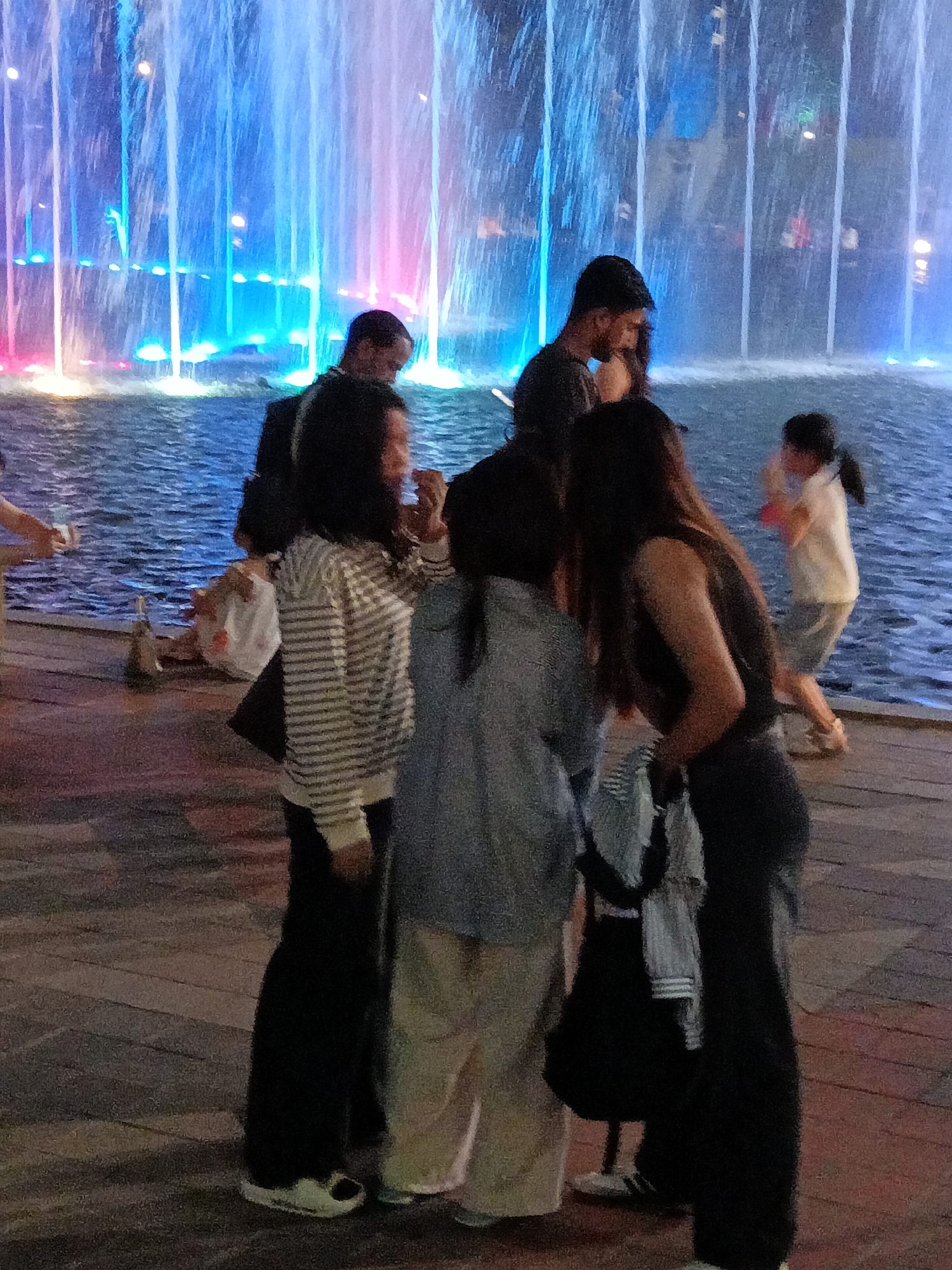 A group of people are gathered near a large, illuminated fountain in Kuala Lumpur, Malaysia. The fountain displays vibrant blue, pink, and white lights as water streams upward. In the foreground, several individuals are interacting. On the left, a person in a striped long-sleeved shirt and black pants stands with their back to the camera. To their right, a person in a gray shirt and light-colored pants is partially obscured. Further to the right, a person in a black sleeveless top and dark pants is visible, interacting with the person in the gray shirt. In the background, to the right, a child in a white shirt and gray shorts is captured in motion, appearing to dance or play near the fountain. The ground is paved with wooden planks. The overall scene suggests a public gathering or a leisurely outing at the fountain.