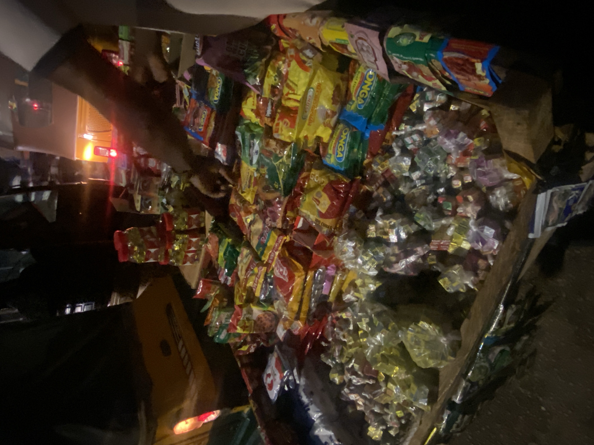 A wooden stand is laden with an assortment of bagged snacks and candies, including several packages of "ONGA" seasoning. A person is standing to the left of the stand, possibly interacting with it. The background appears dark, suggesting it is nighttime. A vehicle with lit headlights and taillights is visible in the background. The location is possibly in the city of Bukuru, Nigeria.