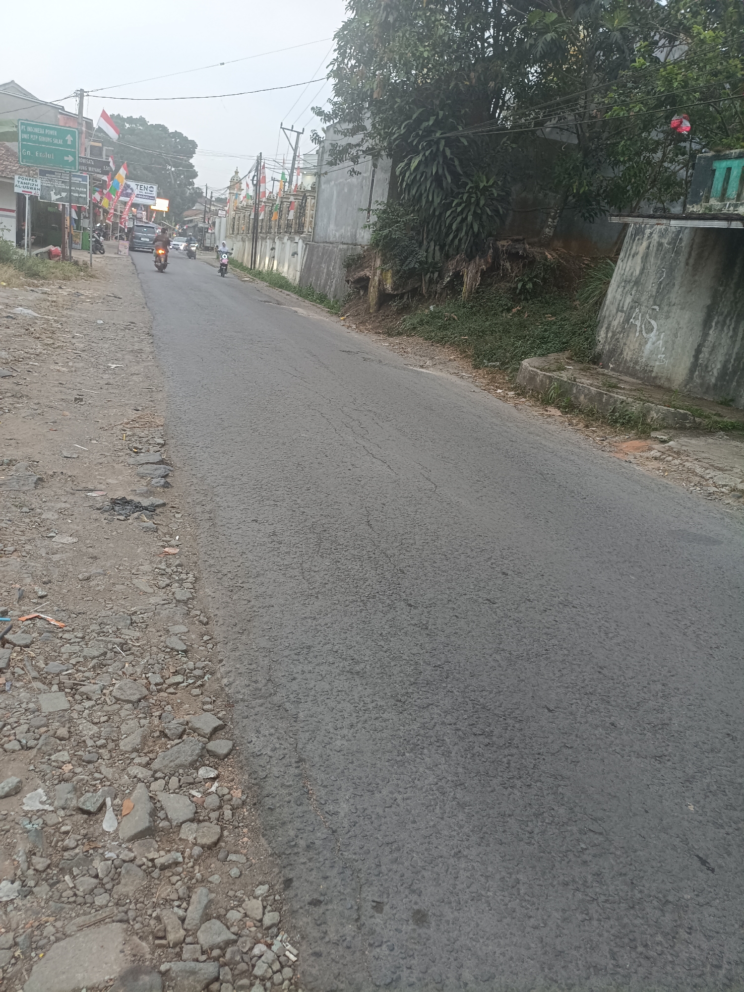 The image depicts a street scene in Berekah, Indonesia, characterized by a paved road with a gravel shoulder on the left. The road curves slightly to the right and recedes into the distance. The shoulder is strewn with rocks, debris, and some litter. Along the right side of the road, there is a raised concrete barrier with some graffiti. Vegetation, including trees and overgrown grass, grows along the embankment behind the barrier.
Several vehicles are visible on the road, including motorcycles and a car. The motorcycles appear to be in motion. On the left side of the road, there are buildings and various signs. One prominent green sign indicates directions to "PT. INDONESIA POWER UNIT PLTP GUNUNG SALAK" and "GN. Endul." Another sign points to "PARIWISATA SUNGAI WAYANG" (Sungai Wayang Tourism). Numerous Indonesian flags are displayed along the roadside, many of them on poles. A large banner for "TENC" is also visible. The sky is overcast, suggesting potentially hazy or foggy conditions. The overall impression is of a rural or semi-urban setting with ongoing activity.