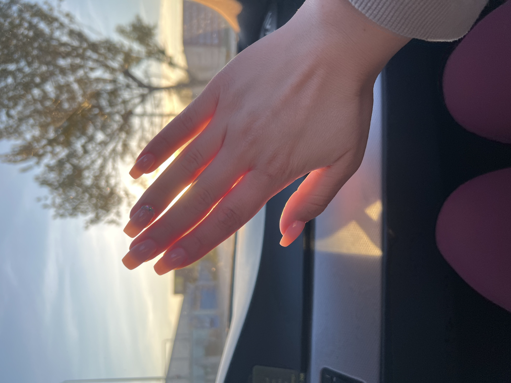 The image displays a close-up of a person's left hand, bathed in warm, golden sunlight. The hand, likely female, is relaxed with fingers slightly spread. The nails are manicured, featuring a translucent peachy-pink polish, and the ring finger's nail is adorned with small, sparkling embellishments. The light catches the edges of the hand, creating a soft glow.
The scene is set inside a vehicle, with elements of the car's interior, such as a dashboard and window frame, visible in the lower and right portions of the image. On the far right, out-of-focus shapes of dark pink or purple fabric, possibly clothing, can be seen.
Beyond the window, a blurred outdoor environment unfolds. A prominent tree with numerous bare or sparsely leaved branches is silhouetted against a bright, hazy sky. The quality of light, characterized by its golden hue and long shadows, strongly suggests either sunrise or sunset. This tranquil moment, capturing the serene beauty of the golden hour, takes place in Plovdiv, Bulgaria. No discernible text is visible in the image.