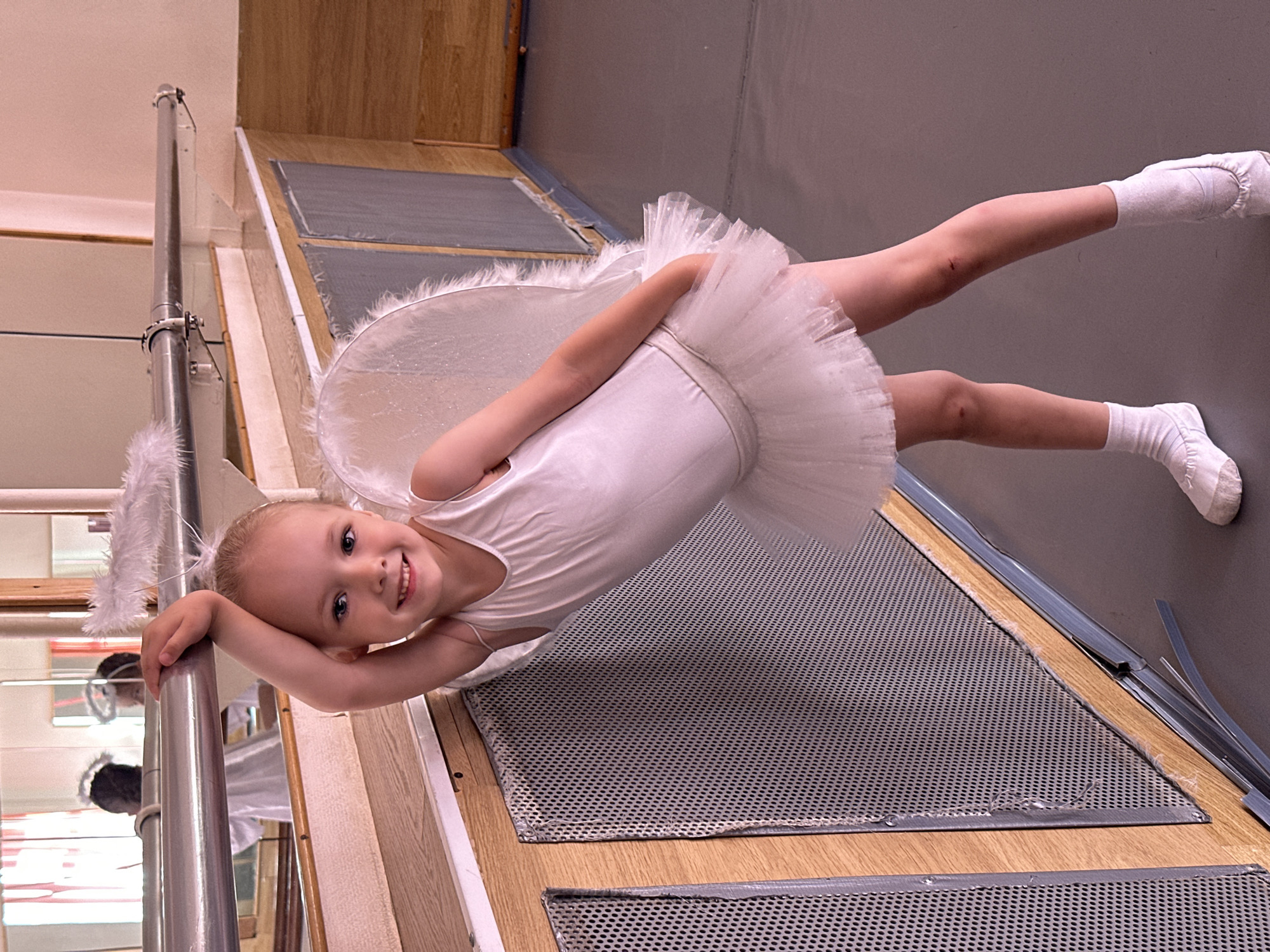 A young girl, likely a child, is posed in what appears to be a dance studio or similar training space in the city of Chaikovsky, Russia. She is wearing a white leotard, a white tutu, white socks, and what seem to be small white angel wings. She is holding onto a ballet barre. The background includes wooden floors, metallic grates, a mirror, and the barre system. The girl is smiling at the camera.