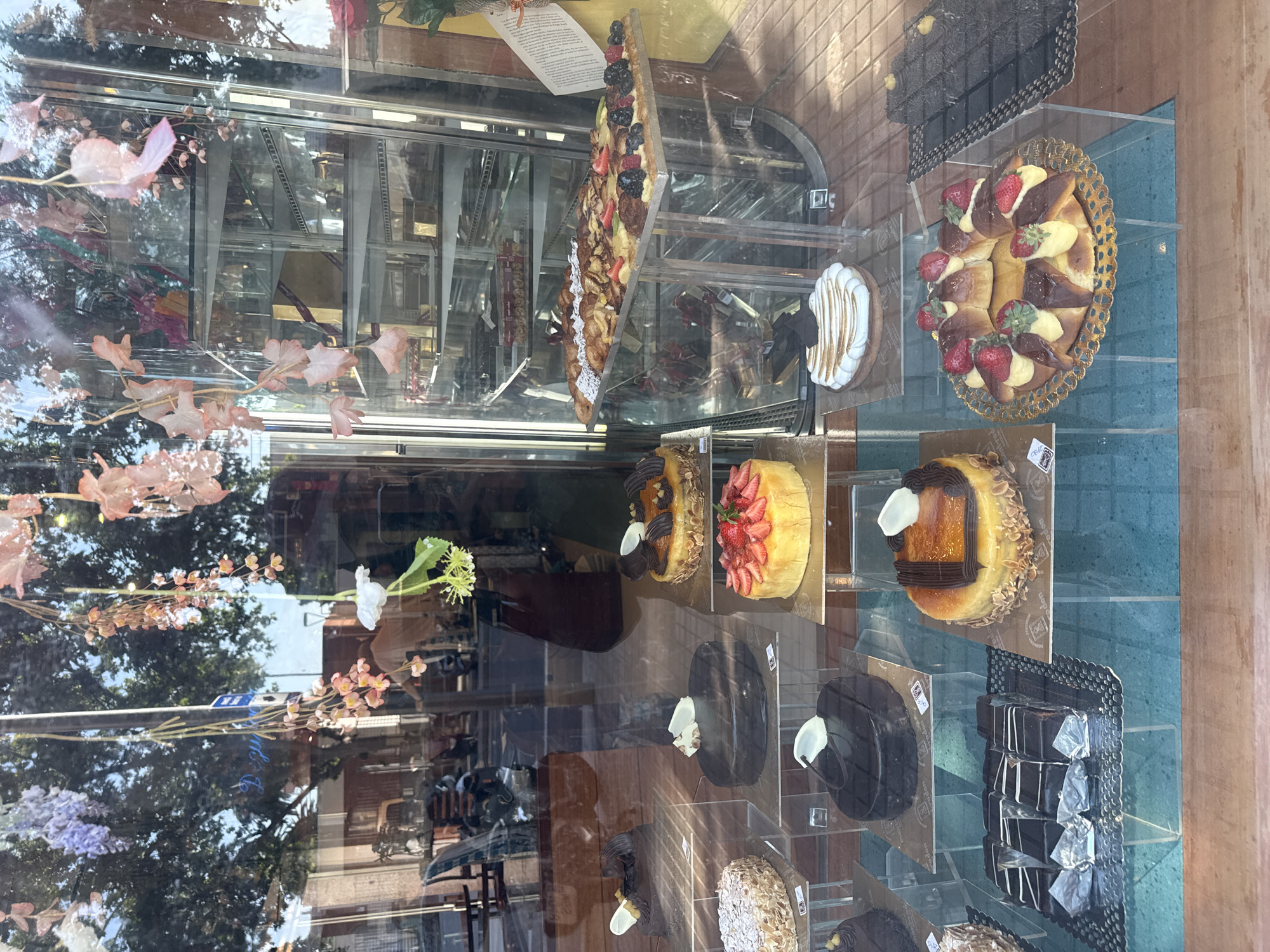 The image depicts a bakery storefront in Barcelona, Spain, displaying a variety of pastries and cakes in glass display cases. Reflected in the glass are trees, buildings, and street elements, giving context to the location. The pastries include fruit tarts, chocolate cakes, and cream-filled pastries. The setting appears to be an upscale bakery.