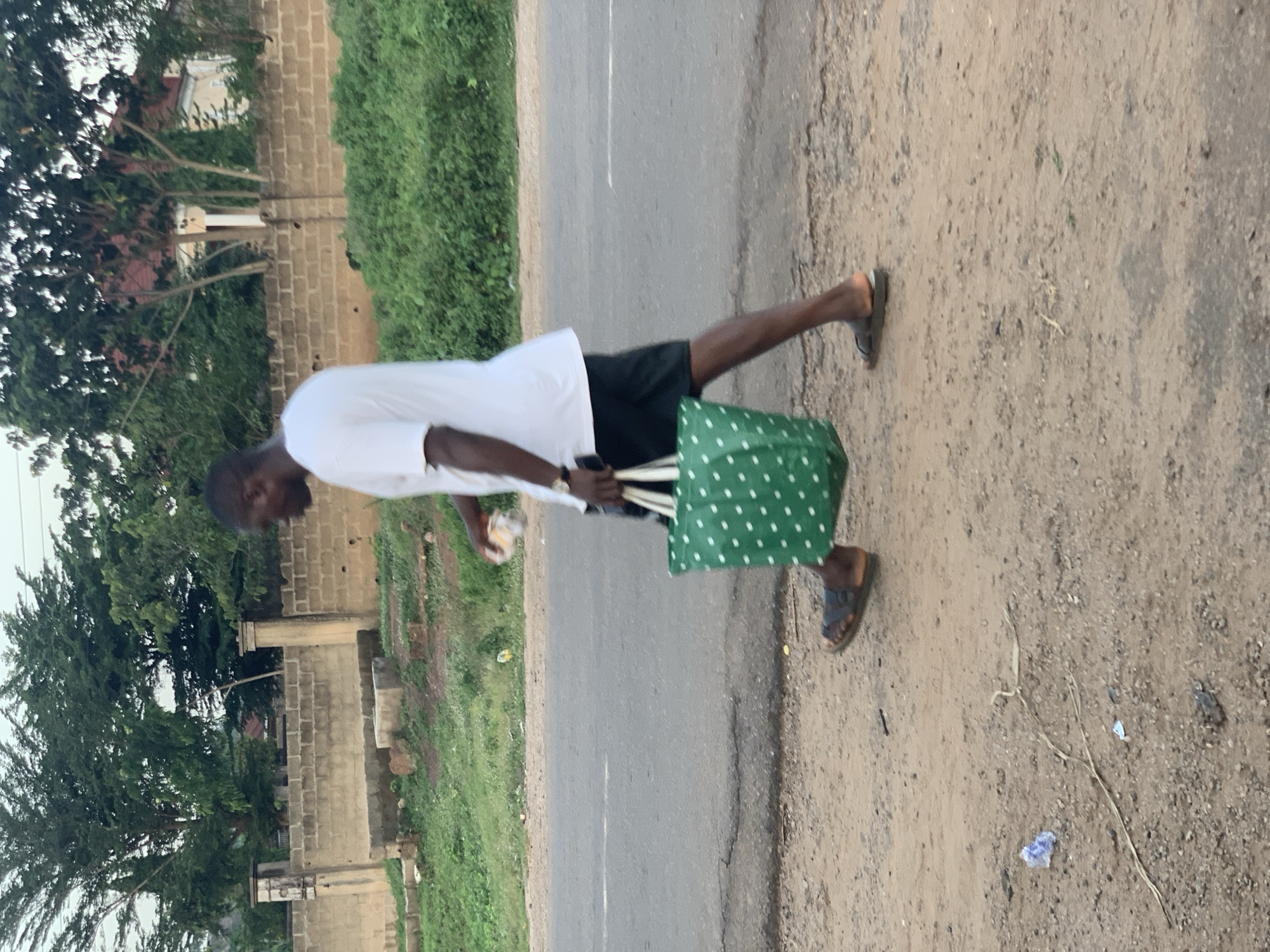 A man is walking along the side of a paved road in Gbeleko, Nigeria. The man is wearing a white shirt, black shorts, and sandals. He is carrying a green bag with white polka dots in his right hand and a white object in his left. The road is cracked and has some debris on the side. On one side of the road is green vegetation and a partially constructed building made of cinder blocks. On the other side is a dirt shoulder with some dry vegetation. The sky is overcast.