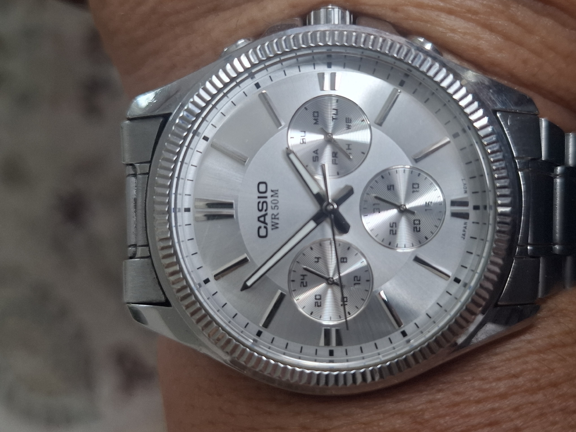 The image is a close-up, eye-level shot of a Casio wristwatch on a person's wrist. The watch has a silver-colored metal band and case, and a silver dial with silver hour markers and hands. There are three sub-dials on the watch face, displaying 24-hour time, day of the week, and a chronograph function. The watch also has "CASIO WR 50M" printed on the dial, indicating its water resistance. The background is blurred and appears to be an indoor setting, possibly a home, with a textured wall or fabric visible. The image does not provide any information about the location in Almaty, Kazakhstan.