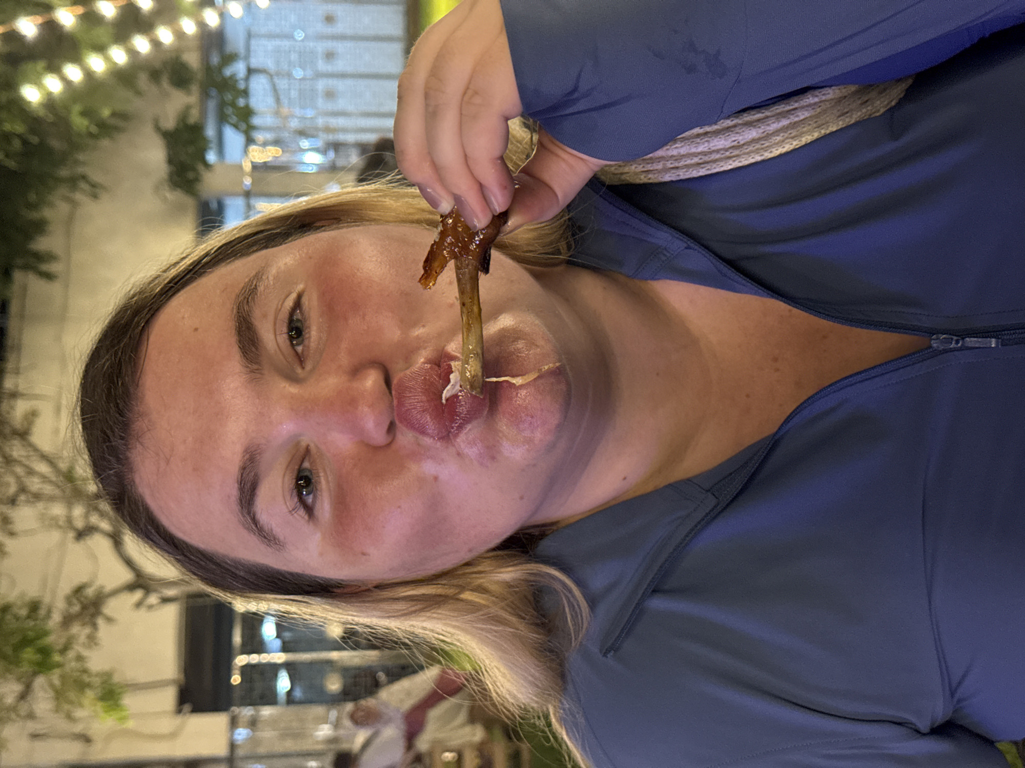 In Phuket City Municipality, Thailand, a medium shot captures a blonde woman with fair skin and rosy cheeks, wearing a blue zippered jacket, as she brings a chicken wing to her mouth. Her lips are pursed as she prepares to eat. In the background, blurred lights suggest an outdoor evening setting, possibly a market or street food stall, with trees and foliage visible above.
