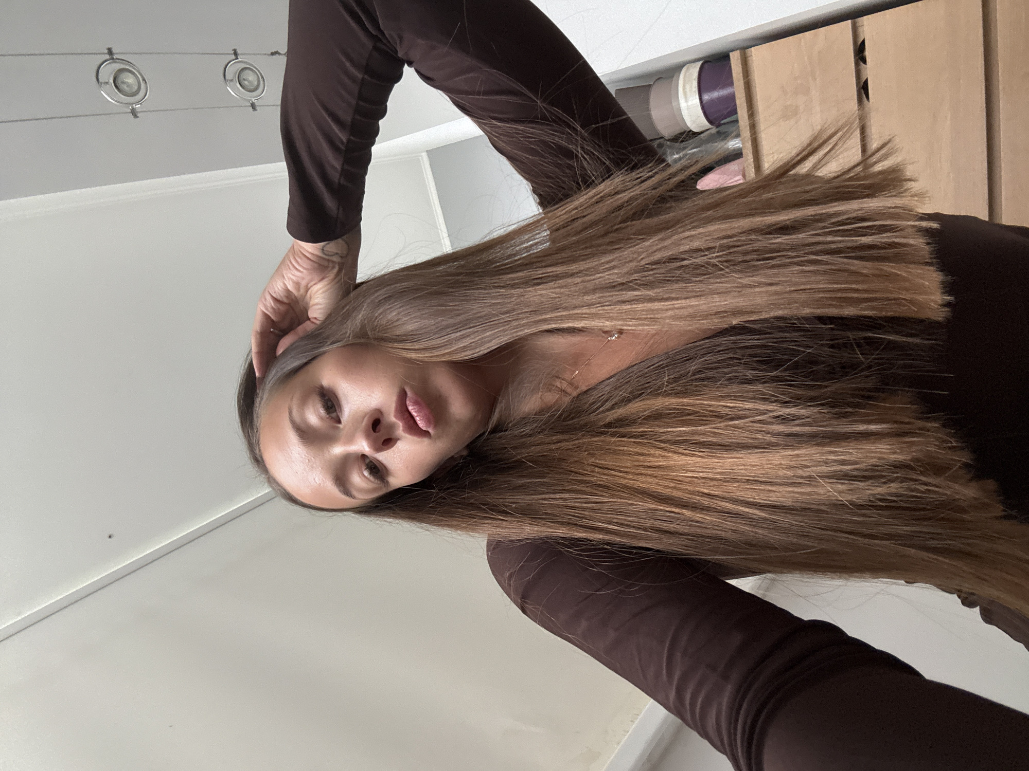 A woman with long brown hair and brown clothing is looking down at the camera. Her left hand is touching her head. In the background, there is a white ceiling with track lighting, a white wall, and a partially visible wooden shelf. The location is not provided and cannot be inferred from the image.