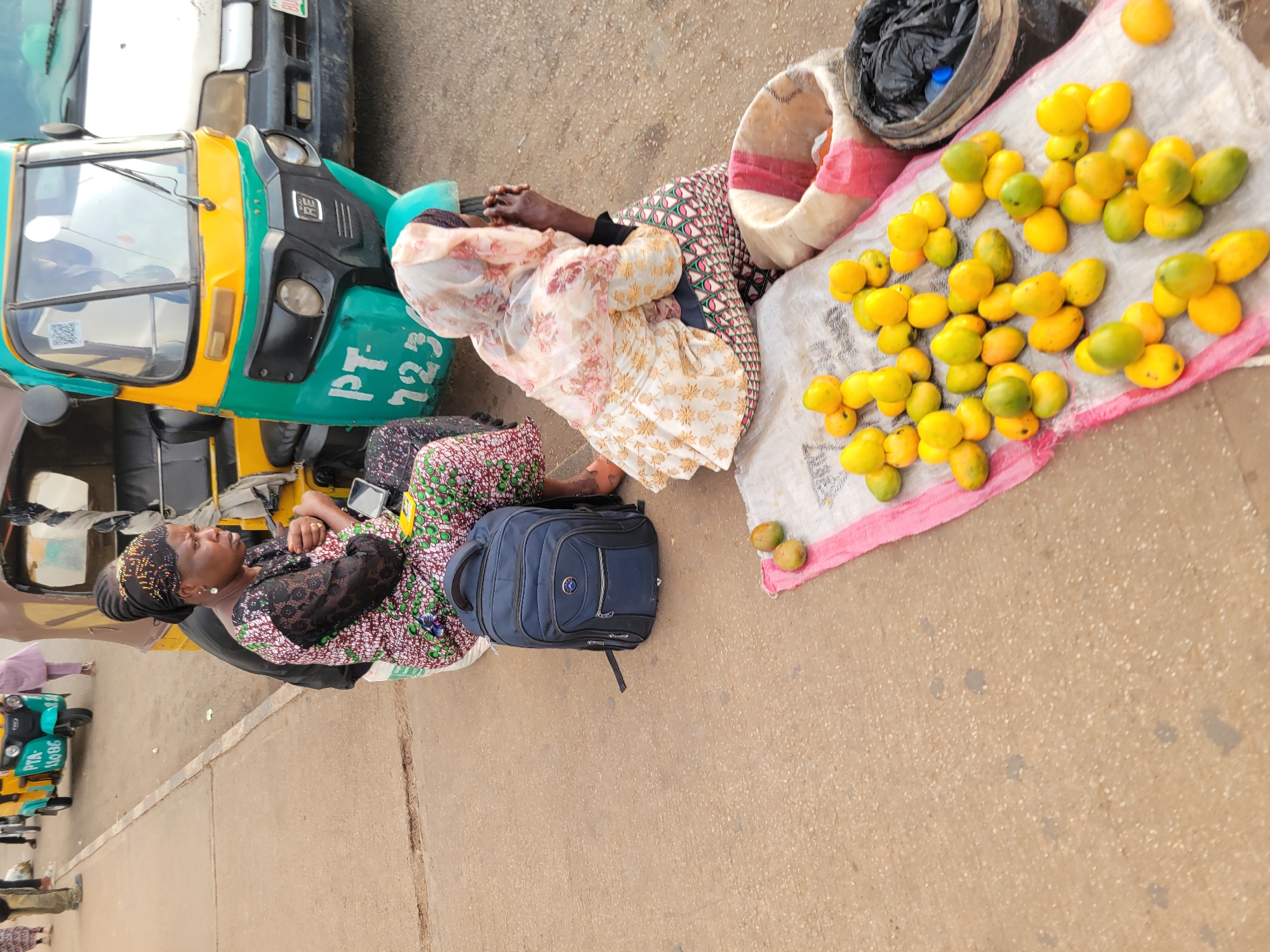 In Barakin Lerawi, Nigeria, a street scene unfolds with two women engaged in commerce. One woman, seated, wears a patterned dress and a headscarf, her gaze directed to the right. Beside her rests a blue backpack. The second woman, also seated, is clad in a patterned headscarf and a floral dress, her hands clasped in front of her. A pile of ripe mangoes is displayed on a white cloth spread on the ground, some in small clusters, others scattered. To the right, a woven basket holds a black plastic bag and possibly more produce. In the background, a bright yellow and green auto-rickshaw, identified by "PT 723" on its side, and a white vehicle suggest a busy transportation hub. Other individuals, partially visible, contribute to the bustling atmosphere of the Nigerian marketplace.