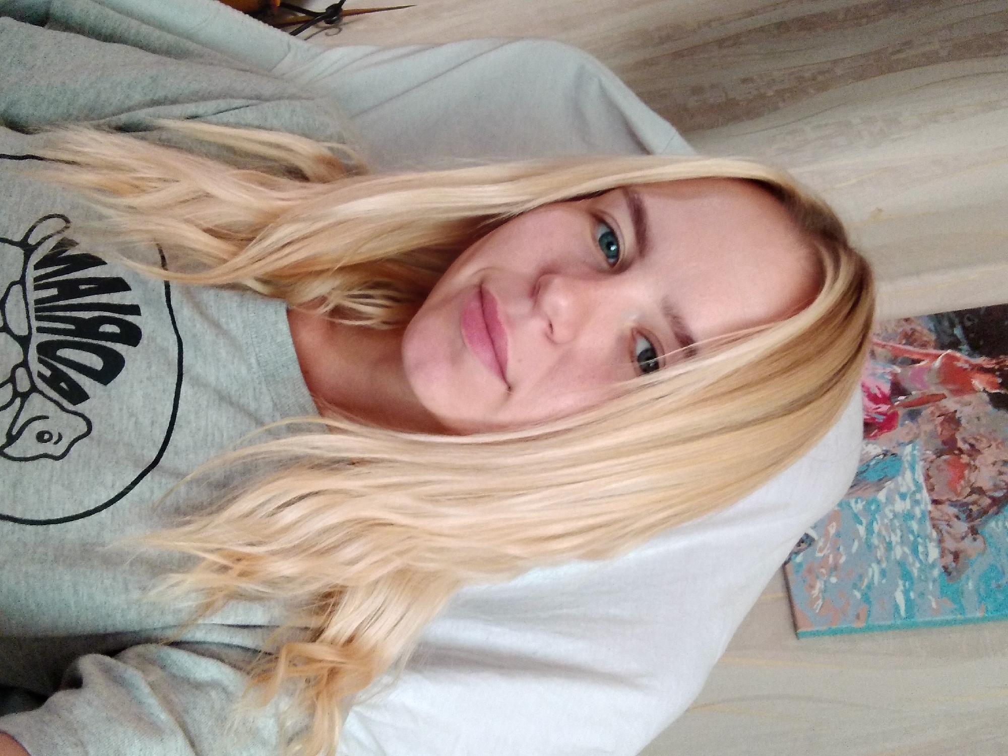 A young woman with blonde hair and blue eyes is photographed from the chest up, sitting in a chair. She is wearing a gray sweatshirt with a black graphic design. A painting is visible on the wall behind her. The image does not provide enough context to determine the location within Radisne, Ukraine.