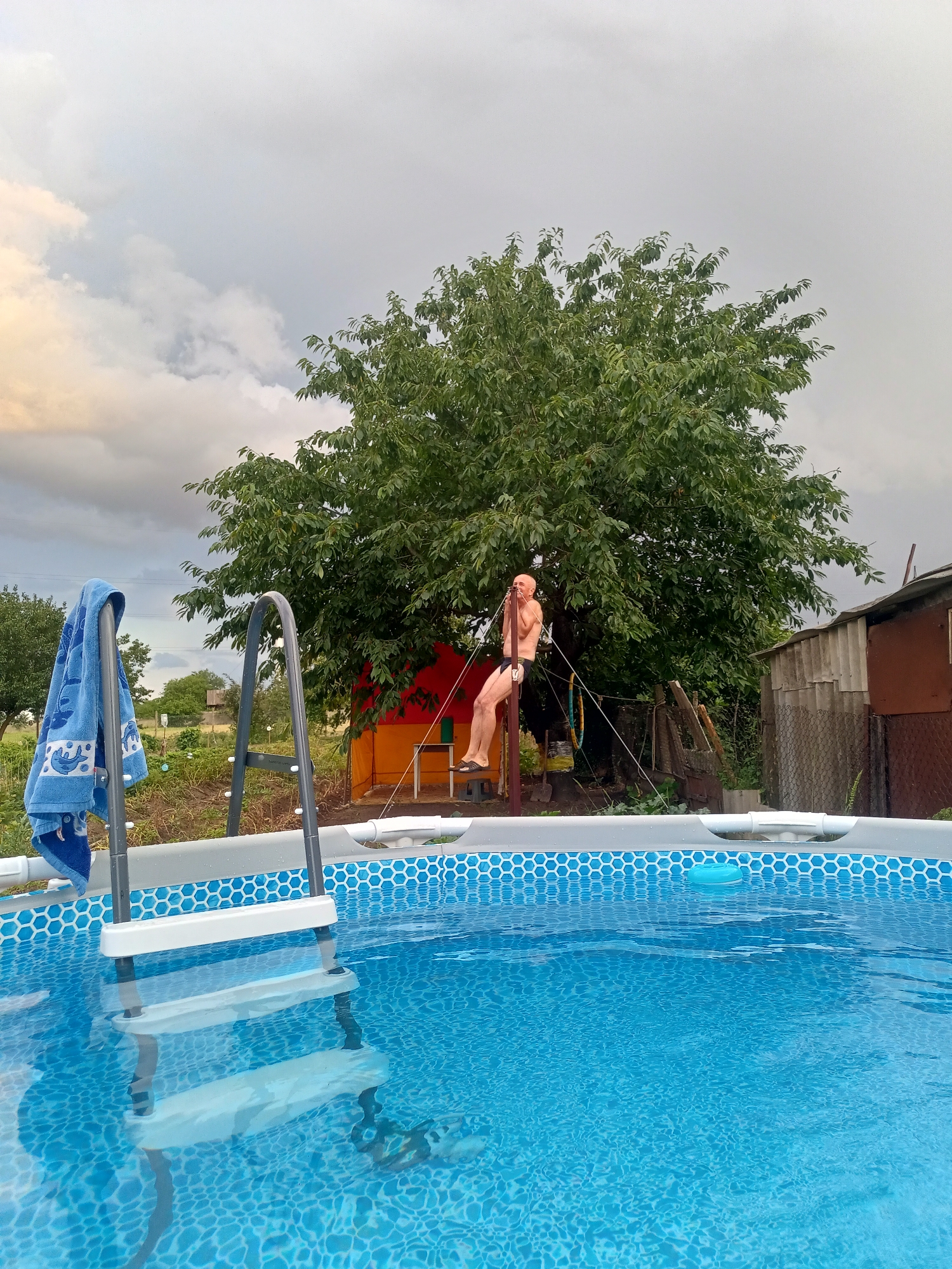 An outdoor swimming pool is shown in Salhany, Ukraine, with a man hanging from a rope swing attached to a large tree. The pool is filled with blue water and has a ladder leading into it. A blue towel is hanging on the ladder. The man is wearing black swim trunks. Behind the tree, there is a red and orange structure and a dilapidated shed. The sky is cloudy.