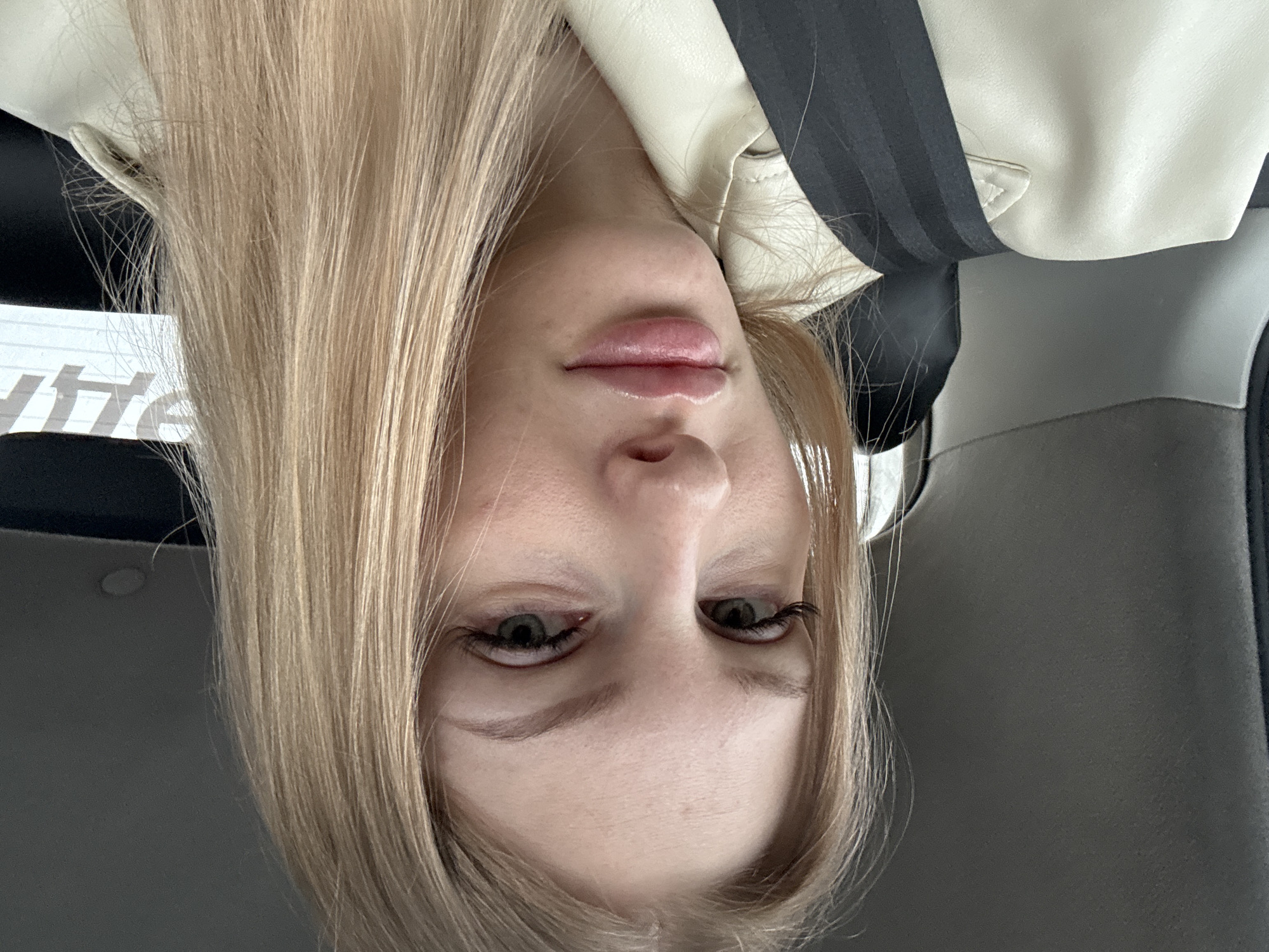 A blonde-haired woman is depicted in an upside-down selfie. She is wearing a cream-colored jacket with a black and gray strap across her chest, possibly a seatbelt. The background appears to be the interior of a vehicle, with gray upholstery visible. There is no evidence in the image to confirm the location is the city of Perm, Russia.
