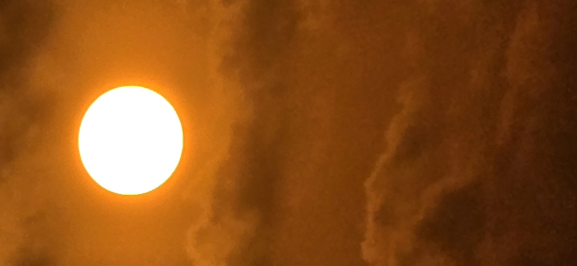 The image shows a bright, white sun, nearly fully illuminated and centrally located in the frame. It is set against a backdrop of hazy, orange-toned clouds, suggesting either a sunrise or sunset. The lower portion of the image reveals further layers of darker, amorphous clouds, creating a sense of depth and atmosphere. There are no people, buildings, or other discernible man-made structures visible in the image, making it an abstract natural scene. The image does not provide any specific visual cues that would allow for the identification of its location within the city of Bat Yam, Israel.