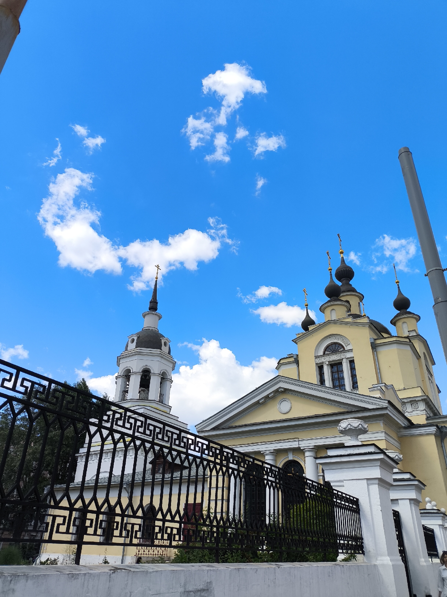 This media file depicts a church and its bell tower against a vibrant blue sky dotted with fluffy white clouds. The scene is shot from a low angle, emphasizing the grandeur of the religious structures. A decorative black metal fence runs across the foreground, partially obscuring a white wall and lush greenery.
The main subject is a yellow church with multiple onion domes topped with crosses. To the left, a white bell tower with a dark dome and a cross also stands prominently. The architecture suggests a historical or traditional religious building. A tall, gray cylindrical object, possibly a utility pole or a part of a building, is visible on the right side of the frame, reaching towards the sky.
The weather appears to be pleasant with a clear blue sky and scattered clouds, indicating it might be daytime, likely late morning or afternoon given the bright lighting. The overall mood is serene and reverent, befitting the subject matter. No people are clearly visible in detail, though a small part of a person in a white shirt can be seen on the far right edge of the image. There is no visible text in the image. The location is unknown.