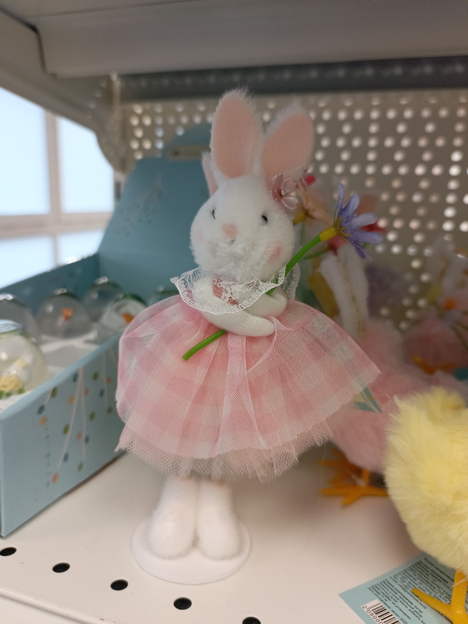 A close-up shot captures a plush white bunny wearing a pink tutu and holding a blue flower. The bunny is positioned on a white shelf next to a light blue box containing glass orbs, and a yellow chick toy. A tag is partly visible near the chick with text in Russian. The background features a white, patterned lattice. The bunny, chick and box are all on a white metal shelf. The tag suggests the items are located in Chaikovsky, Russia.