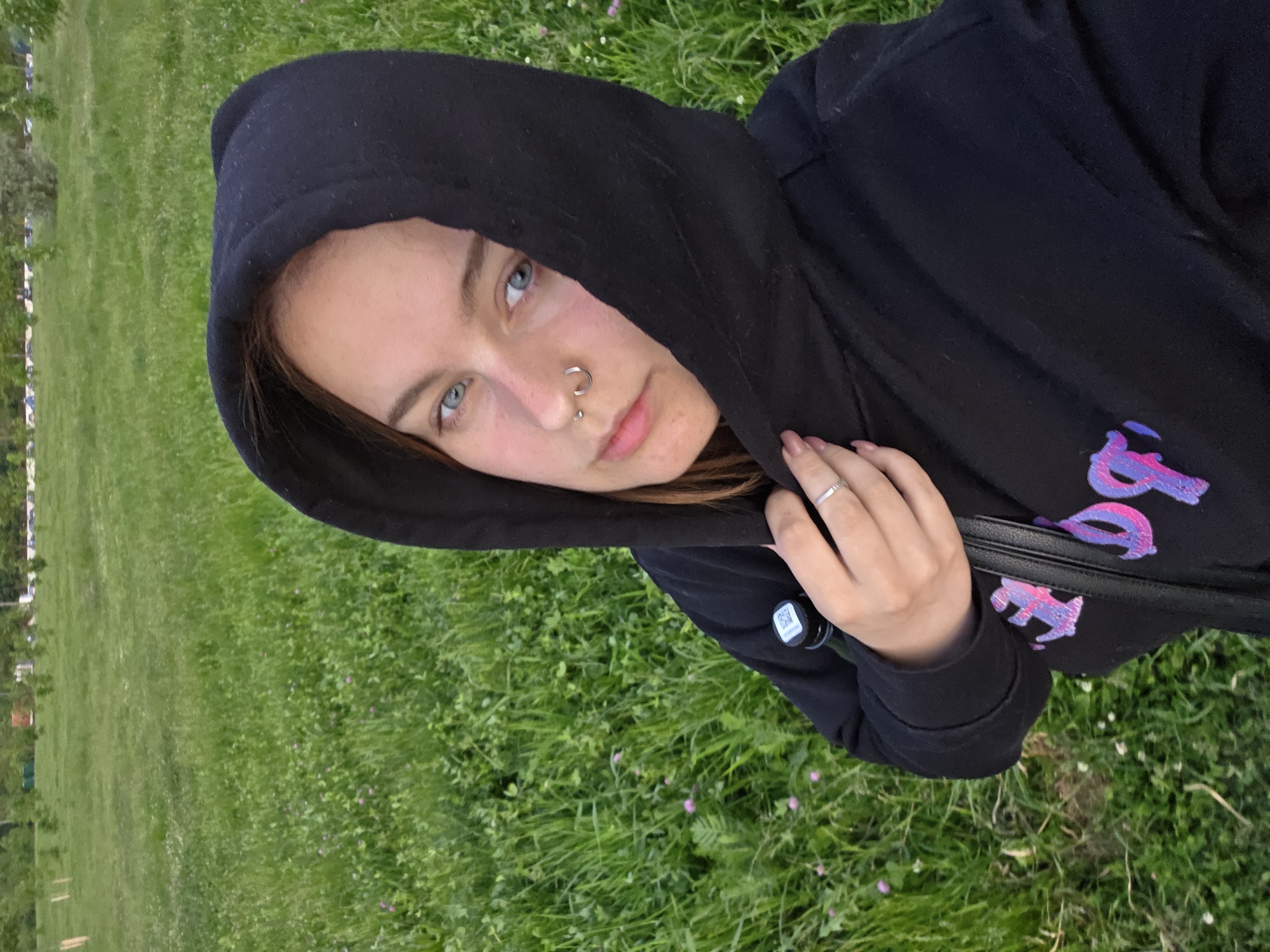 The image depicts a young adult female taking a selfie outdoors in an open grassy field in the city of Unknown, Unknown. The subject has blue eyes, a fair complexion, and light brown hair partially visible beneath the black hood of her sweatshirt. She has a silver septum ring and a silver stud in her left nostril. Her expression is neutral, and she looks directly into the camera. Her left hand, wearing a silver ring on the ring finger, lightly holds the neckline of her black hoodie. The hoodie features a partially visible graphic in pink and purple on the chest. A dark-colored bottle with a black and white cap is visible near her right shoulder. The background consists of a vibrant green lawn with scattered small purple flowers. In the far distance, a line of trees and faint structures are visible under a soft, diffused light.
