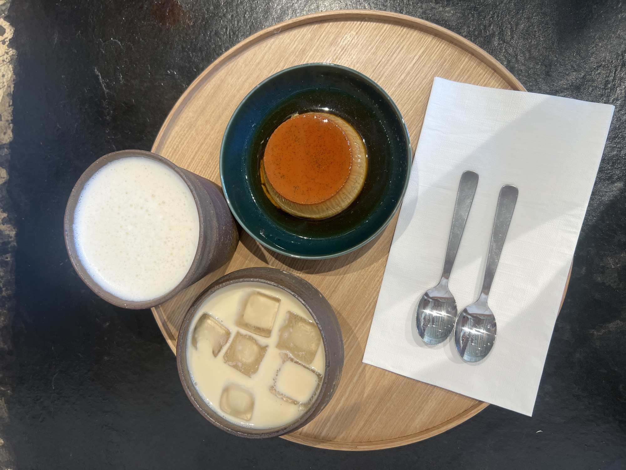 A top-down shot depicts a wooden tray containing two beverages, a dessert, and two spoons resting on a white napkin. The tray sits on a dark, textured surface that could be a table.
On the left, two ceramic cups of different sizes are filled with drinks; the cup above holds a white, foamy liquid, while the one below contains a creamy beverage with ice cubes. Adjacent to the cups is a green bowl containing a flan-like dessert. To the right of the bowl is a folded white napkin, topped with two metal spoons.
The location is unspecified; there is no information to say if it is in New York City or not.