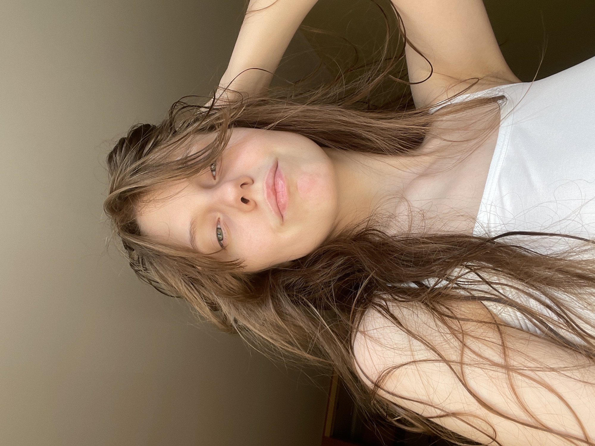 The image is a close-up, low-angle, selfie-style photograph of a young Caucasian female. She has long, brown, wavy hair that appears wet and is somewhat disheveled, framing her face and falling over her shoulders. Her skin is fair with a slight blush on her cheeks. She is wearing a white tank top. Her left hand is raised and resting on the back of her head, with strands of hair cascading around her arm and shoulder. Her expression is neutral, with her eyes looking slightly upwards and to her left. The background is a plain, muted brown, suggesting an indoor environment with diffused lighting, possibly near a window. There is no discernible indication of the city of Ufa, Russia, or any specific location within it. The image is a portrait focusing on the individual.