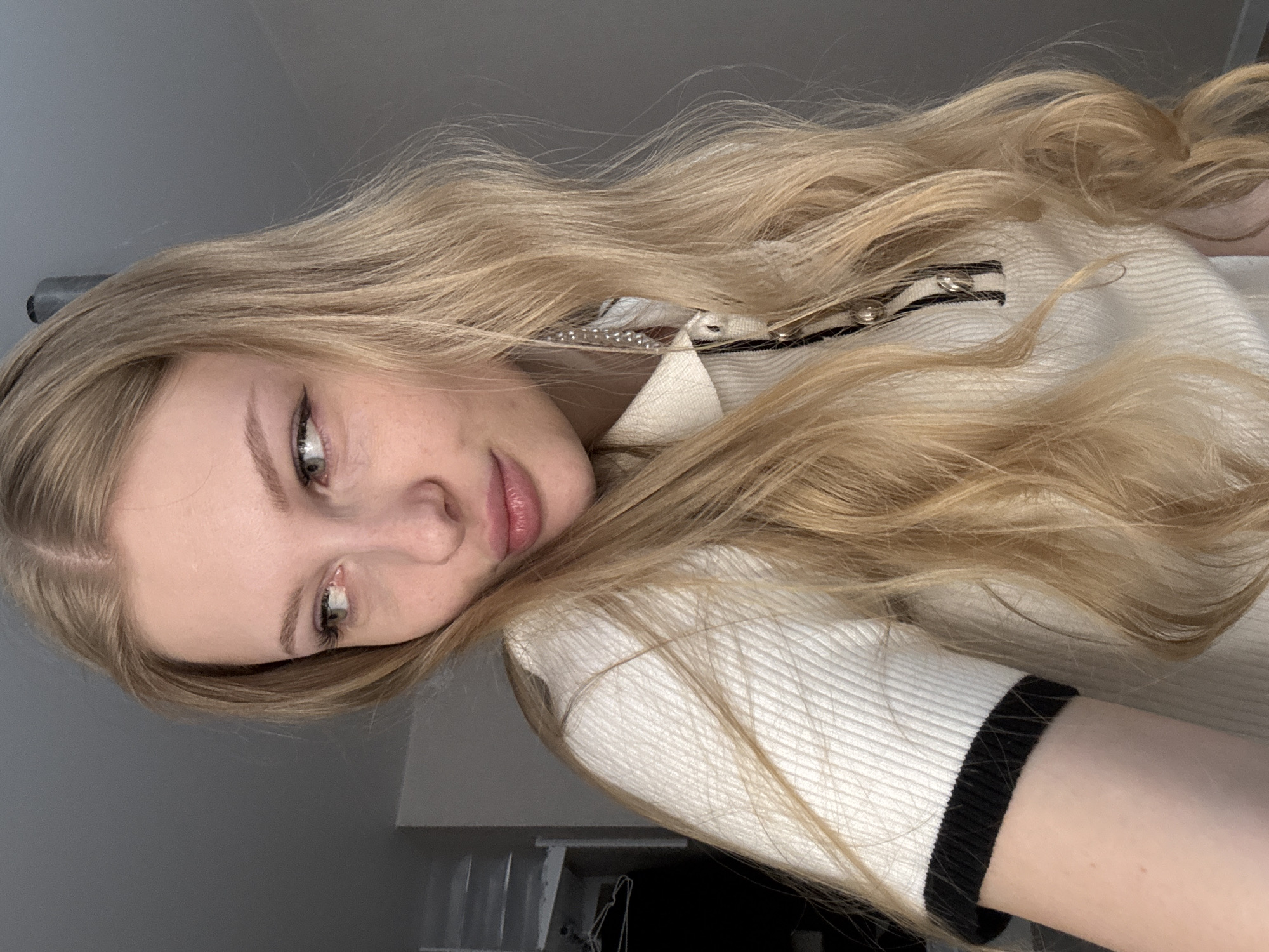 A medium shot captures a young female indoors, featuring long, wavy blonde hair with a central parting. Her eyes are light, accented by dark winged eyeliner, and her lips are full with a light pink tone. She is looking to the viewer's left with a slightly pouting expression. She wears a white, ribbed, short-sleeved polo shirt with a black and white striped placket secured by silver buttons and black trim on the sleeve cuffs. A long, multi-beaded earring is visible on her right ear, partially obscured by her hair. The background is a plain, dark grey wall, with a white shelving unit and a clothes hanger partially visible on the far left, suggesting an interior room. The image contains no identifiable visual cues to place the subject or scene within the city of Perm, Russia.