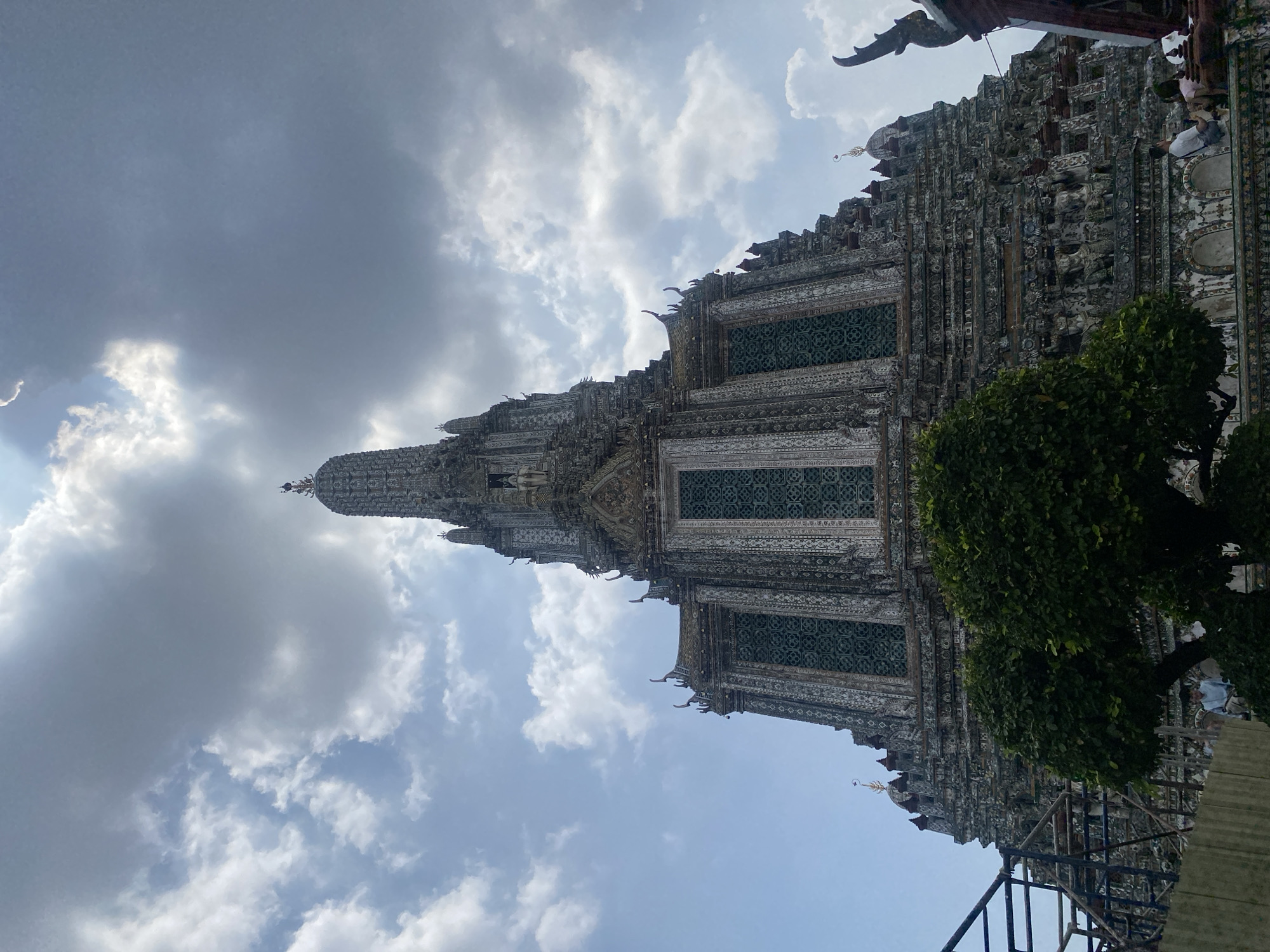 The image shows Wat Arun, a Buddhist temple in Bangkok, Thailand. The temple's central prang (tower) is the focal point, rising prominently against a cloudy sky. The prang is decorated with intricate mosaic patterns. A tree partially obscures the lower right corner of the temple. Construction scaffolding is visible at the base of the temple.