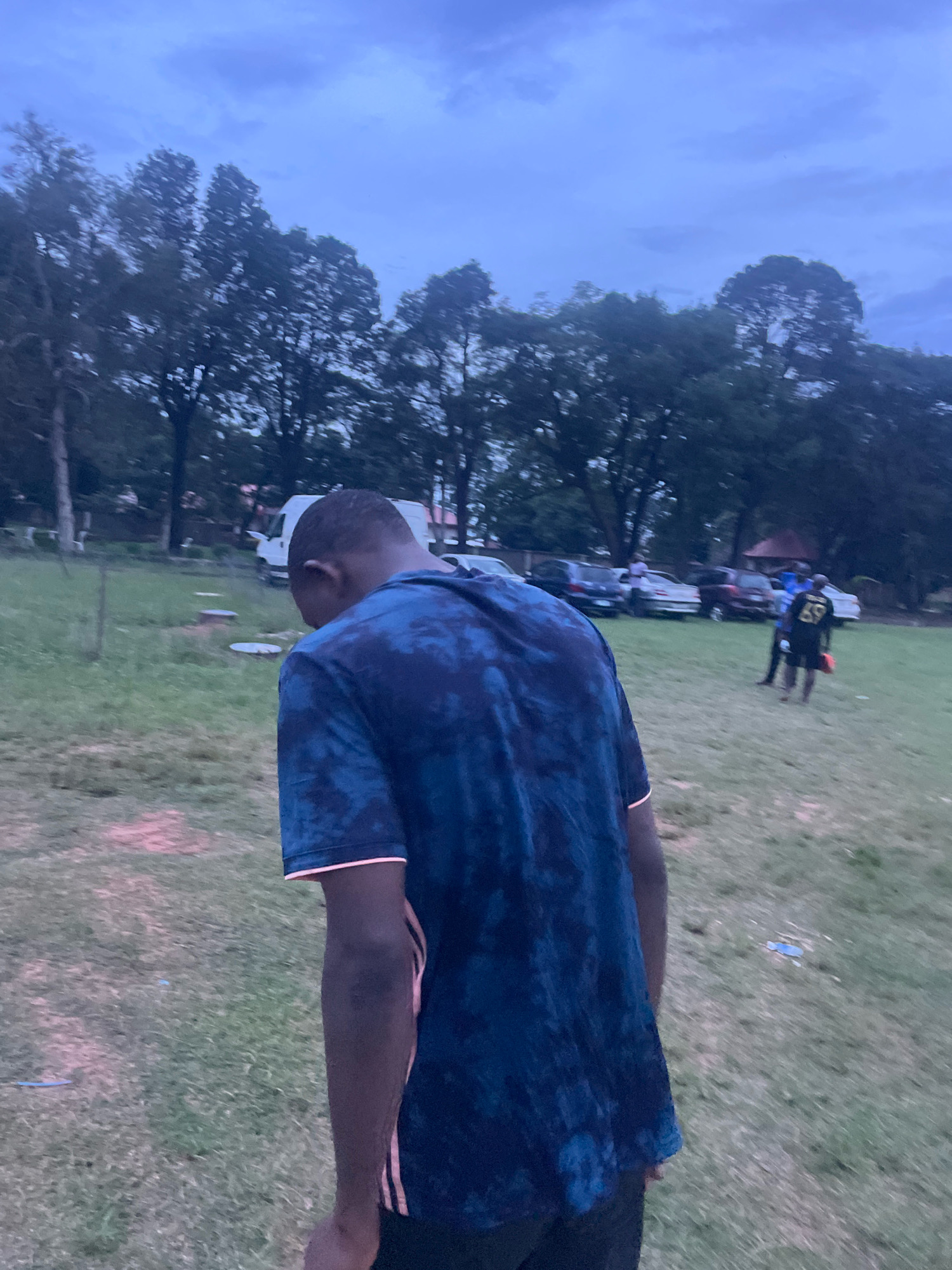 The image depicts an outdoor scene, likely in the city of Jos, Nigeria, under a dusky sky with overcast clouds. In the foreground, a person wearing a dark blue tie-dye t-shirt with pink accents is facing away from the camera, their head down. The person's arm is visible, showing dark skin and pink stripes down the side of the shirt.
The background reveals a grassy area with scattered trees. Several vehicles are parked in the middle ground, including a white van and various cars. In the far background, two individuals are visible; one is wearing a dark shirt with the number '69' on the back and dark shorts, holding a red object, and the other person is wearing a blue shirt. The overall impression is a gathering or event taking place outdoors in a park-like setting, possibly a field adjacent to a road where vehicles are parked. The lighting suggests it is either early morning or late evening.