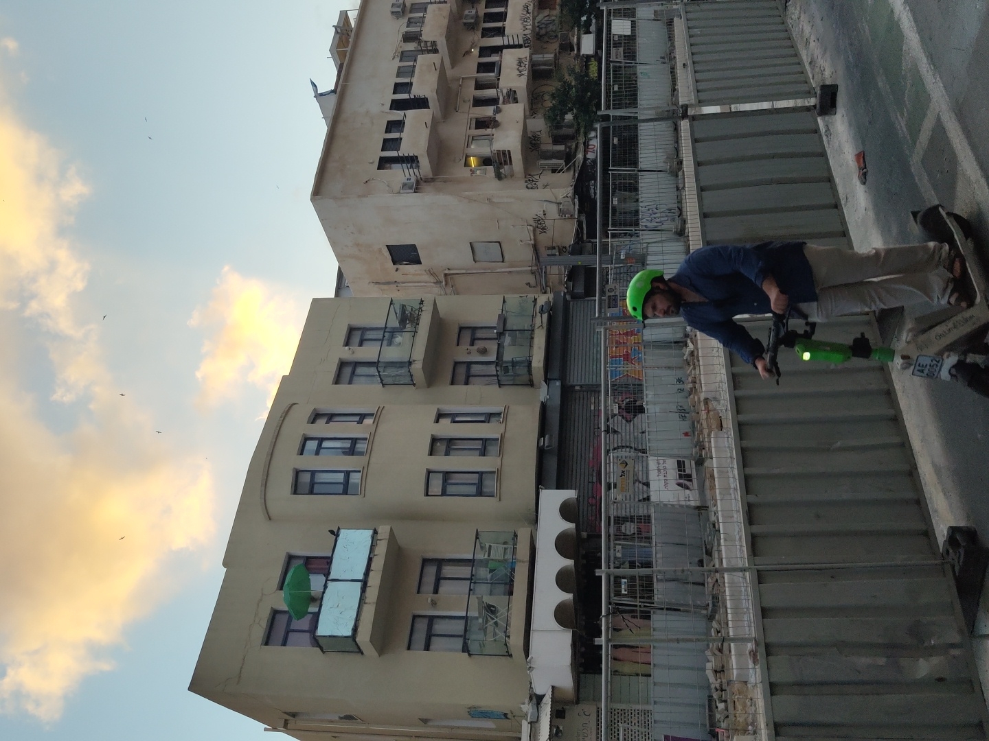 The image depicts a man riding a lime-green electric scooter in Tel Aviv, Israel. He is wearing a blue long-sleeved shirt, beige pants, and a green helmet. The scooter has the license plate "AE 0052". He is riding on a paved street alongside a construction site, which is enclosed by metal fencing. Behind the construction site are two apartment buildings. The building on the left is light beige with a curved facade, featuring several balconies with railings. One balcony has a green umbrella. The building on the right is also beige, with multiple windows and balconies. The sky above is partly cloudy with hints of sunset colors. Birds are visible in the sky. The scene appears to be an urban setting in Tel Aviv, likely during the late afternoon or early evening.