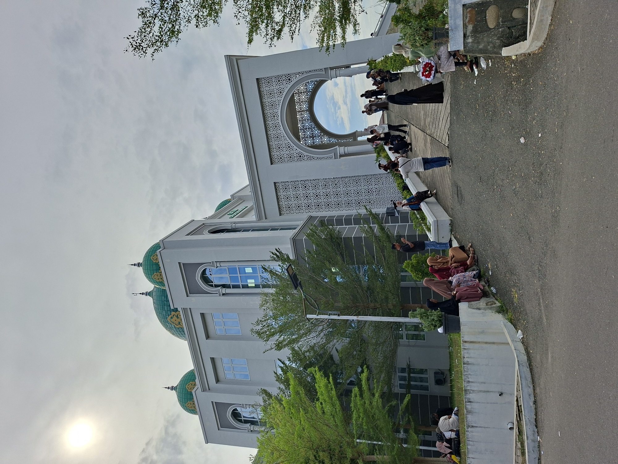 The image captures an outdoor scene featuring a large, grey building with prominent Islamic architectural elements, identifiable as Universitas Al Muslim in Lhokseumawe, Indonesia. The building boasts multiple green domes adorned with golden patterns and features a grand arched entrance with intricate geometric latticework. Numerous windows reflect the bright, partially cloudy sky.
A group of people is gathered near the main entrance, with some standing and others sitting on ledges or steps. Many women are wearing hijabs, and one person holds a red and white floral arrangement, suggesting a special event or gathering at the university. Further along a paved road or pathway to the right, more individuals are seated on a concrete barrier, under the shade of trees. The scene appears social and relaxed, with people engaged in conversations or observing their surroundings.
The time of day is daytime, evidenced by the bright sun peeking through the clouds in the upper left, casting a soft light over the area. Green foliage from trees and bushes is visible around the building and along the path, adding natural elements to the urban setting. The overall atmosphere is calm, possibly indicating a break between classes or a public event at the university. The prominent text "AL MUSLIM" is visible on the building's facade.