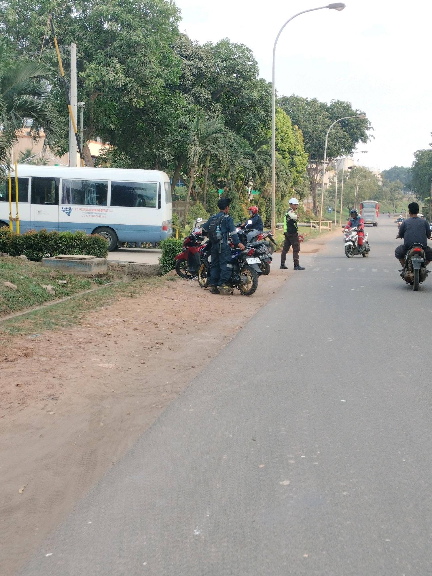 The image depicts a roadside scene in Batam, Indonesia, likely during daylight hours. On the left side of the paved road, a white and blue bus is parked. Adjacent to the bus is a dirt and gravel area with sparse vegetation. Several individuals are gathered near motorcycles on the roadside. One person, wearing a blue uniform, stands with a backpack. Another person, in a red helmet, is seated on a motorcycle. A third person, wearing a white helmet and a high-visibility vest, stands in front of a motorcycle. Further down the road, several other motorcycles are visible, with riders. In the background, there are trees and utility poles with streetlights. The road itself is asphalt, with some white markings.