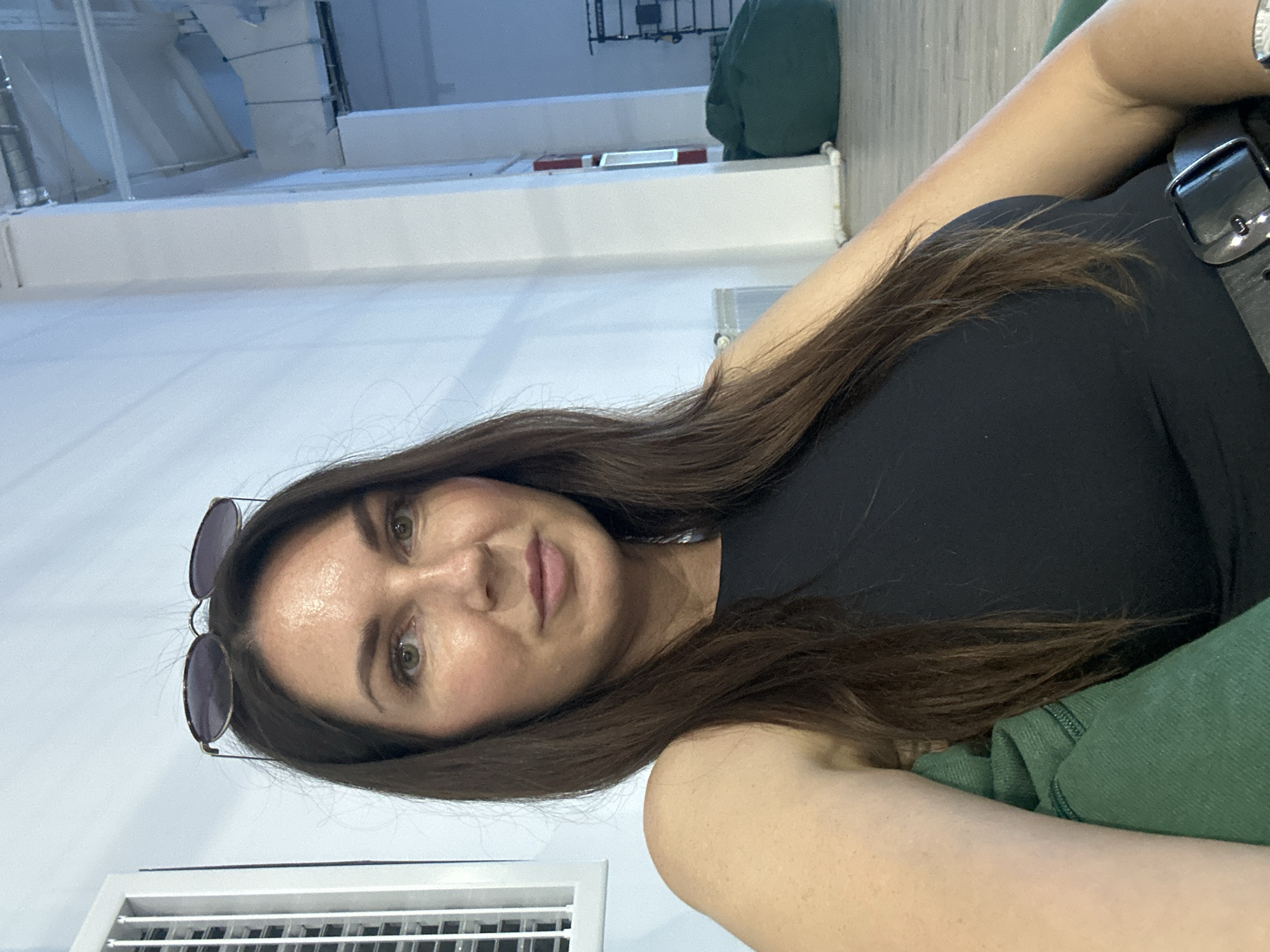 In a well-lit interior space, possibly a gym or studio in Chaikovsky, Russia, a woman with long, dark brown hair is seen from the chest up, seated and looking towards the camera. She is wearing a black sleeveless top and a black belt with a silver buckle. Sunglasses are perched on top of her head. The background features a white wall, a ventilation grate on the left, and to the right, a white pillar, a fire extinguisher, a green beanbag chair, and a wall-mounted exercise apparatus. The floor appears to be light-colored wood.