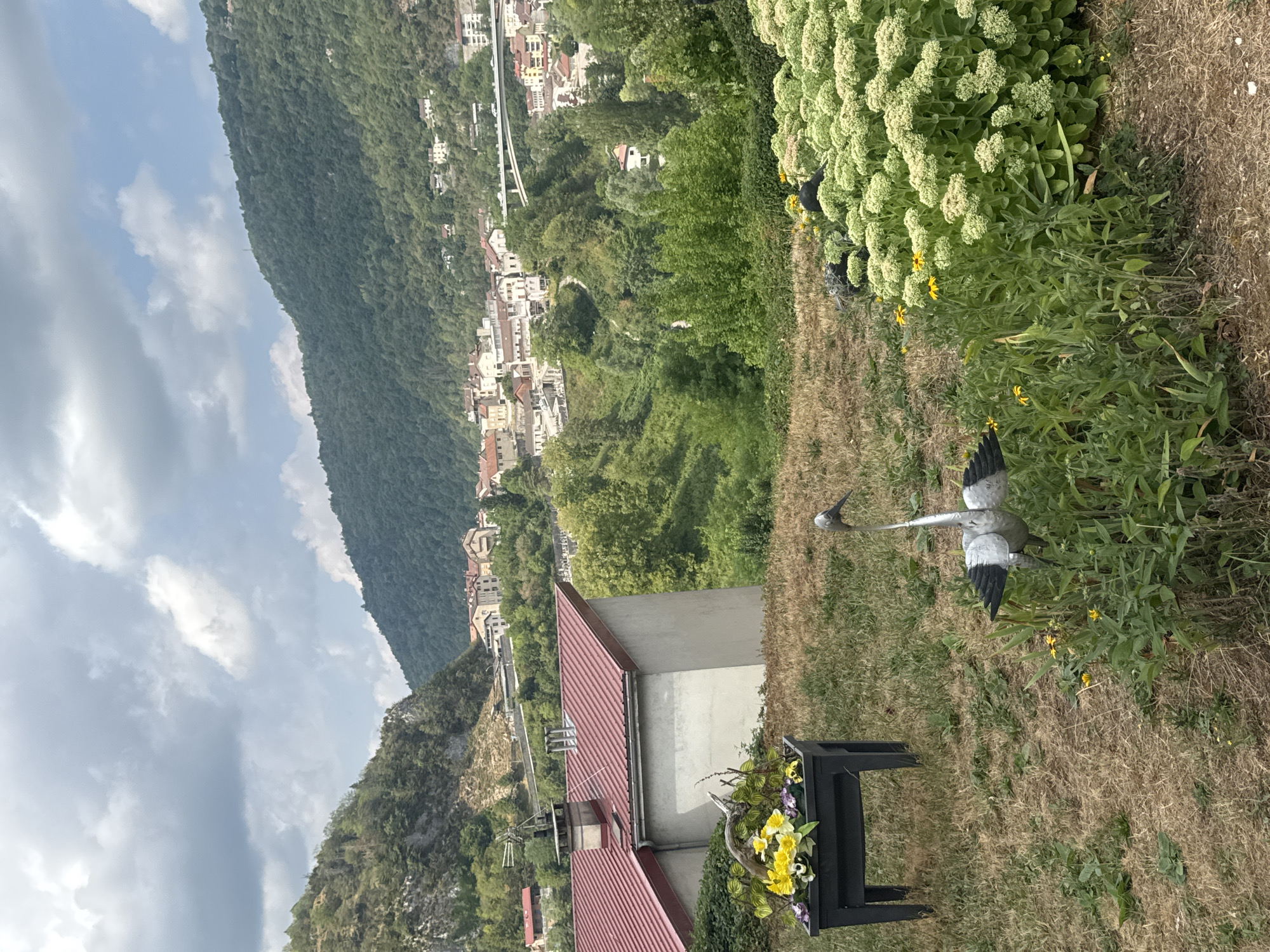 The image captures a scenic view of Saint-Claude, France, with a residential area nestled in a valley surrounded by lush green hills. In the foreground, a dry, grassy area is adorned with a metal sculpture of a bird, resembling a heron or crane. To the left, a dark planter box filled with yellow and purple flowers sits on a patch of dry grass. Behind the planter, a house with a red-tiled roof is visible. The mid-ground features a dense growth of green trees and shrubs, with a town spreading across the hillside. Buildings with terracotta roofs are scattered among the trees, and a white bridge spans across the valley in the distance. The background is dominated by verdant hills under a partly cloudy sky.
