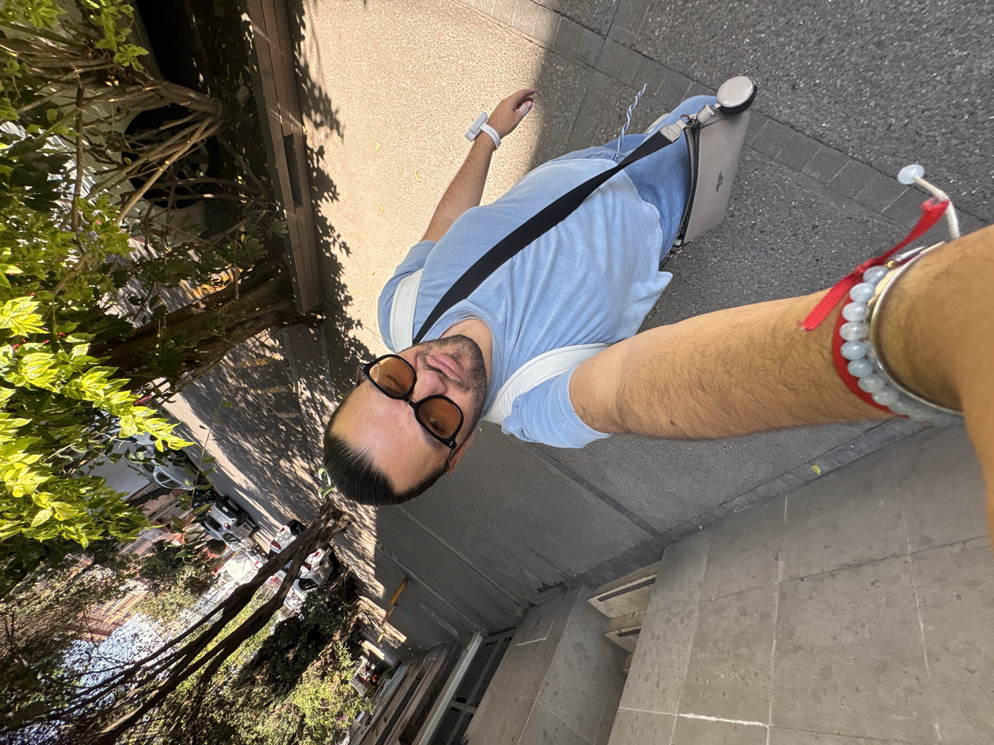A tilted, low-angle selfie captures a man outdoors on a sunny day in an urban environment, identified as Mexico City, Mexico. The man is the central subject, featuring short dark hair, a beard, and amber-tinted sunglasses. He wears a light blue t-shirt and white straps, possibly from a backpack. A gray messenger bag or laptop case with a faint white Apple logo and a silver circular attachment hangs across his chest, secured by a black strap. His left wrist displays a white smartwatch, while his right arm is adorned with several bracelets, including a red string, light blue beaded ones, and a silver-toned piece.
The setting appears to be a city sidewalk or street, with light-colored concrete paving visible. Lush green foliage and mature trees line the area, casting strong, distinct shadows across the ground, indicating bright daylight. In the background, partially obscured by trees, parked vehicles such as a white sedan and a darker car are visible on the street. Multi-story buildings, typical of an urban landscape, stand beyond the trees. The man appears calm, capturing a casual moment during his time outdoors. No other explicit text is discernible.
